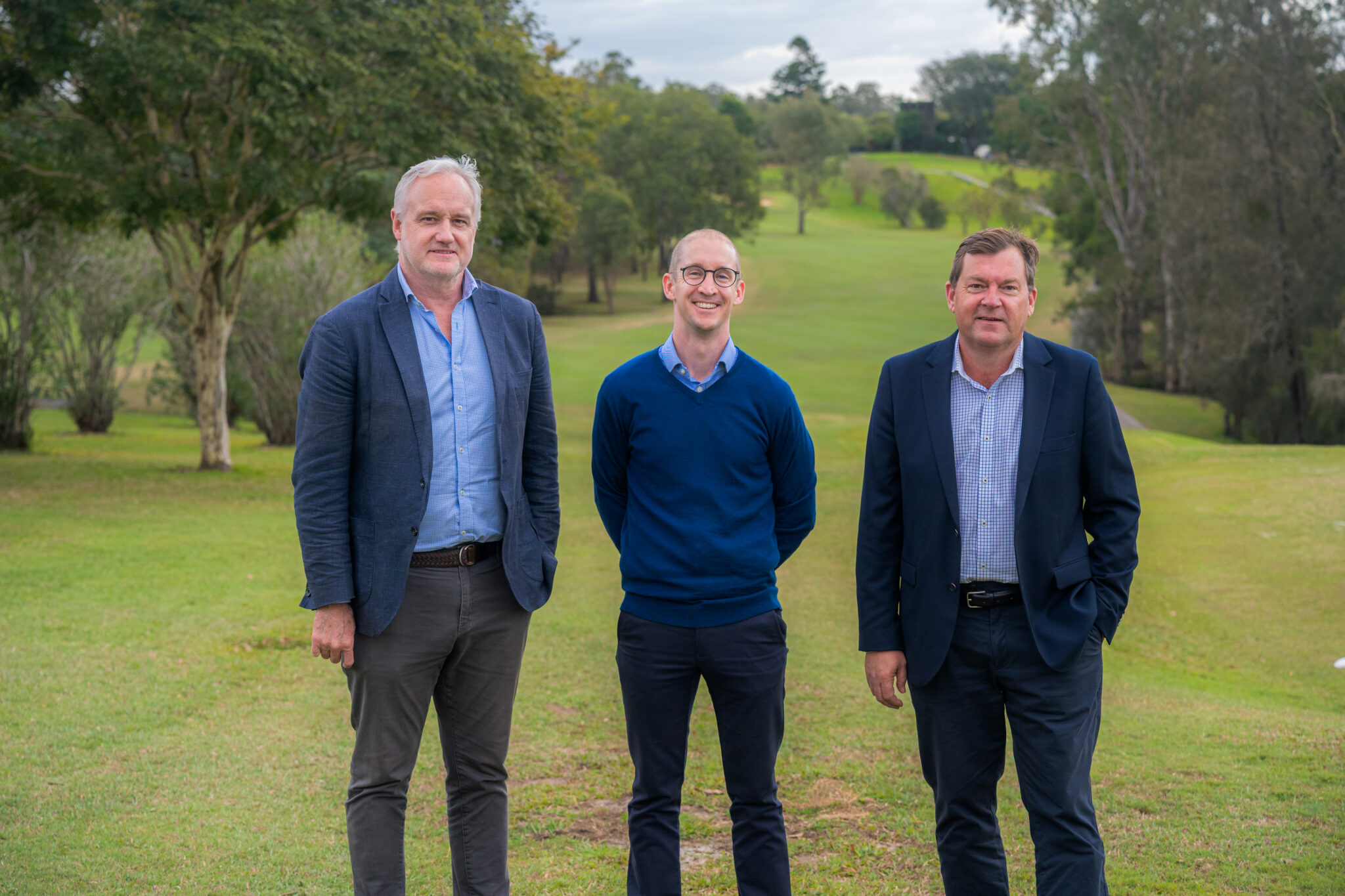 Three men stand side by side on a grassy field in front of pet friendly retirement villages, with trees and rolling hills in the background. All three are dressed in business-casual attire, with jackets and collared shirts.