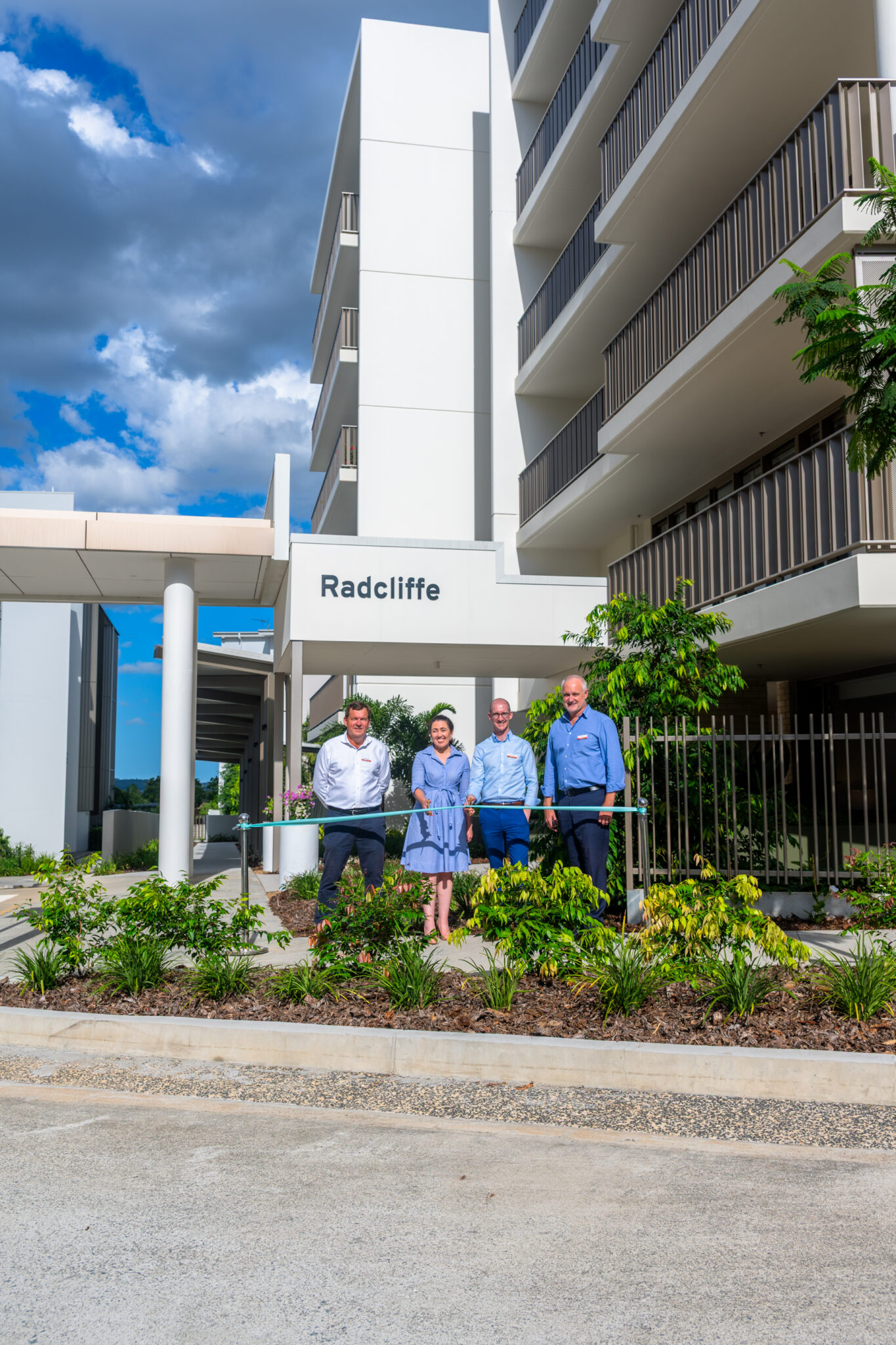 Four people stand in front of a modern apartment building labeled "Radcliffe," one of the area's top pet friendly retirement villages, with landscaped greenery in the foreground and partly cloudy skies above.