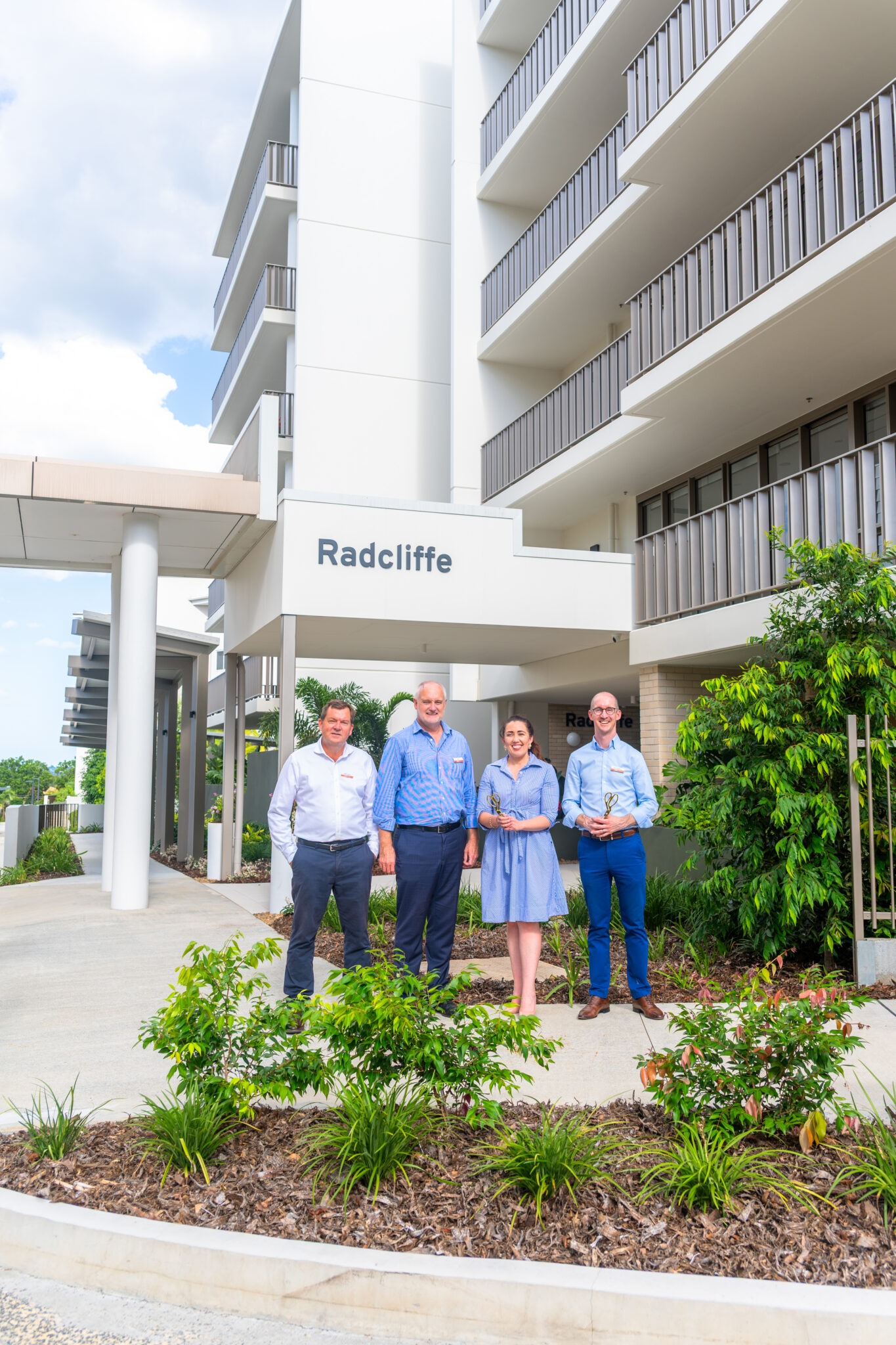 Four residents of Kingsford Terrace stand in front of a modern building with balconies and a "Radcliffe" sign. They are outdoors, surrounded by greenery and landscaping under a partly cloudy sky.