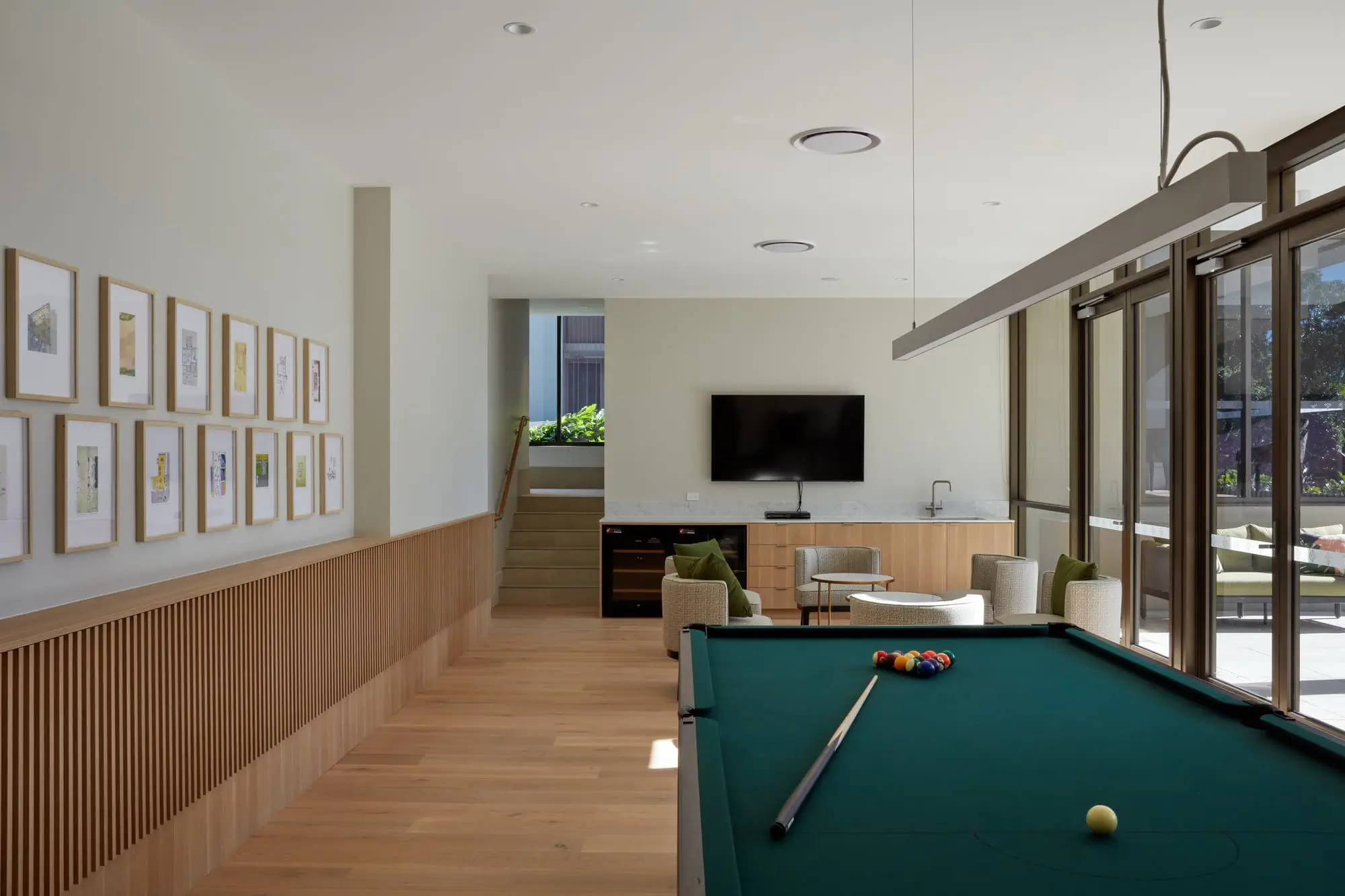 Modern game room with a pool table in the center, cue stick and balls arranged on it, wall-mounted TV, two armchairs, large windows letting in sunlight, and framed pictures on a light-colored wall.