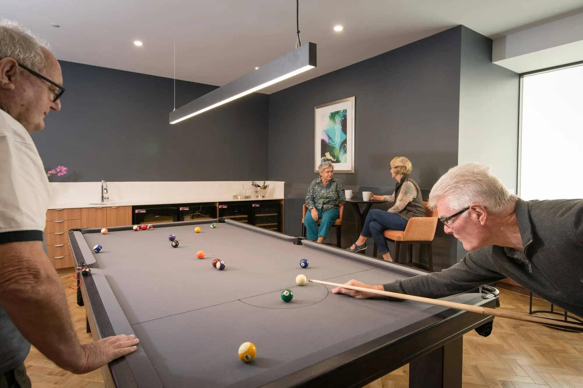 Four older adults are in a modern room; two men are playing pool while two women sit and chat in the background. The space has dark walls, art on the wall, and a long light fixture above the pool table.