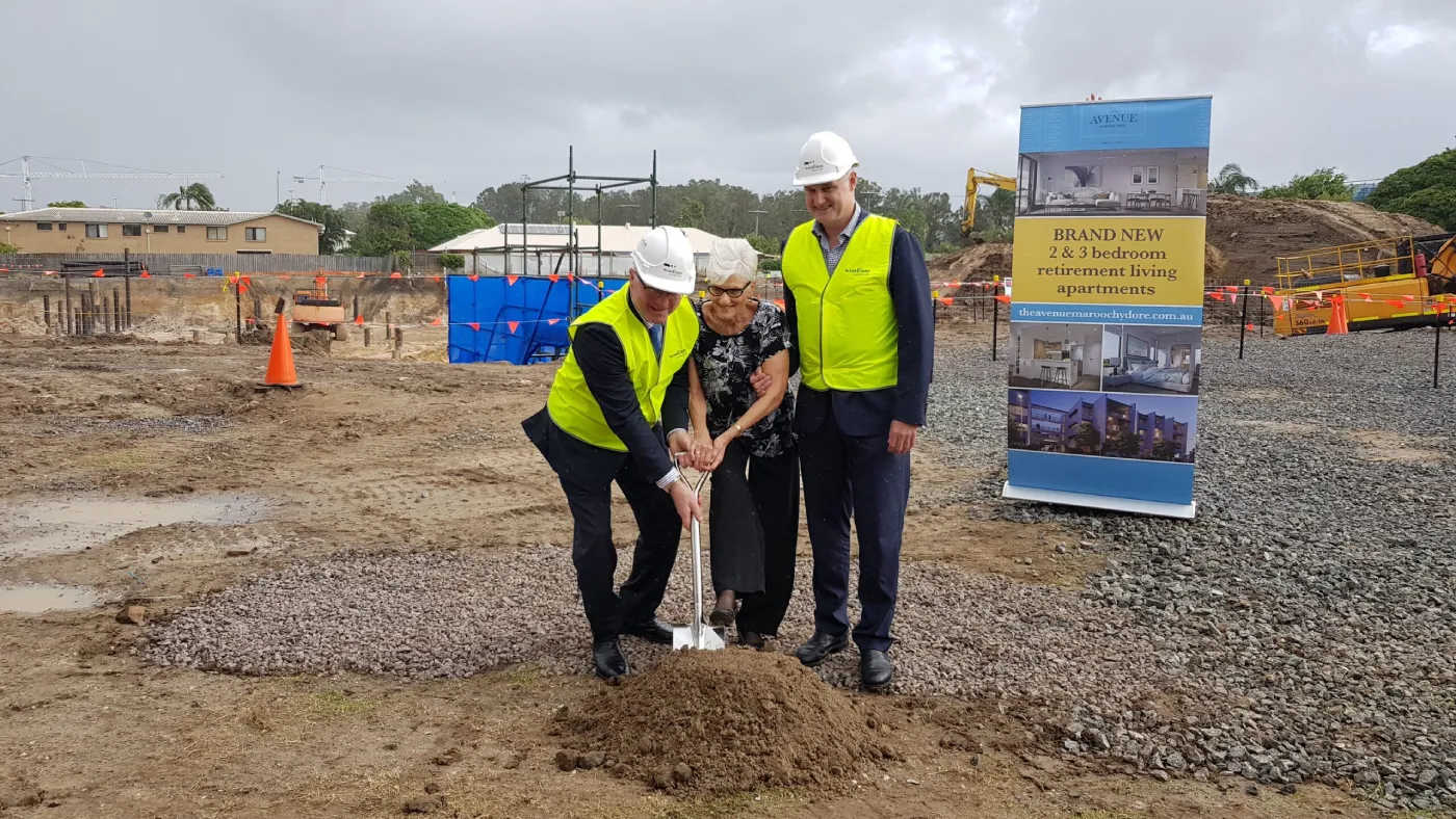 Three people in safety vests and hard hats participate in a groundbreaking ceremony at a construction site, holding a shovel together. A sign nearby advertises brand new retirement living apartments.