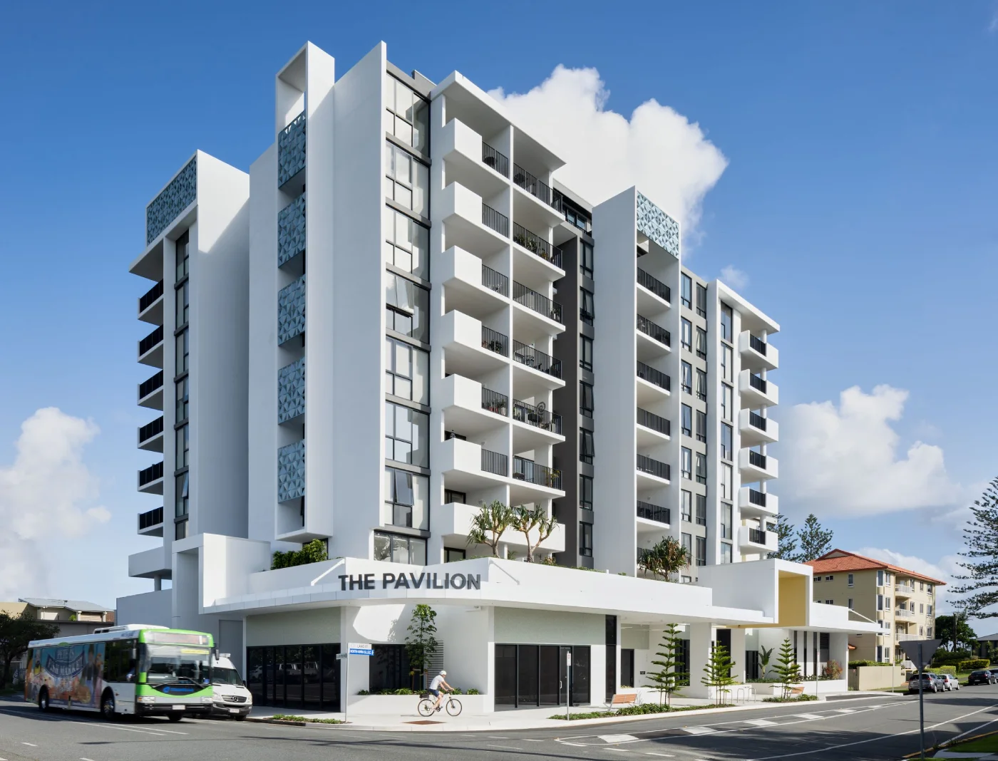 A modern white multi-story apartment building named "The Pavilion" on a sunny day, with balconies, a bus passing by, and a person riding a bicycle on the street corner.