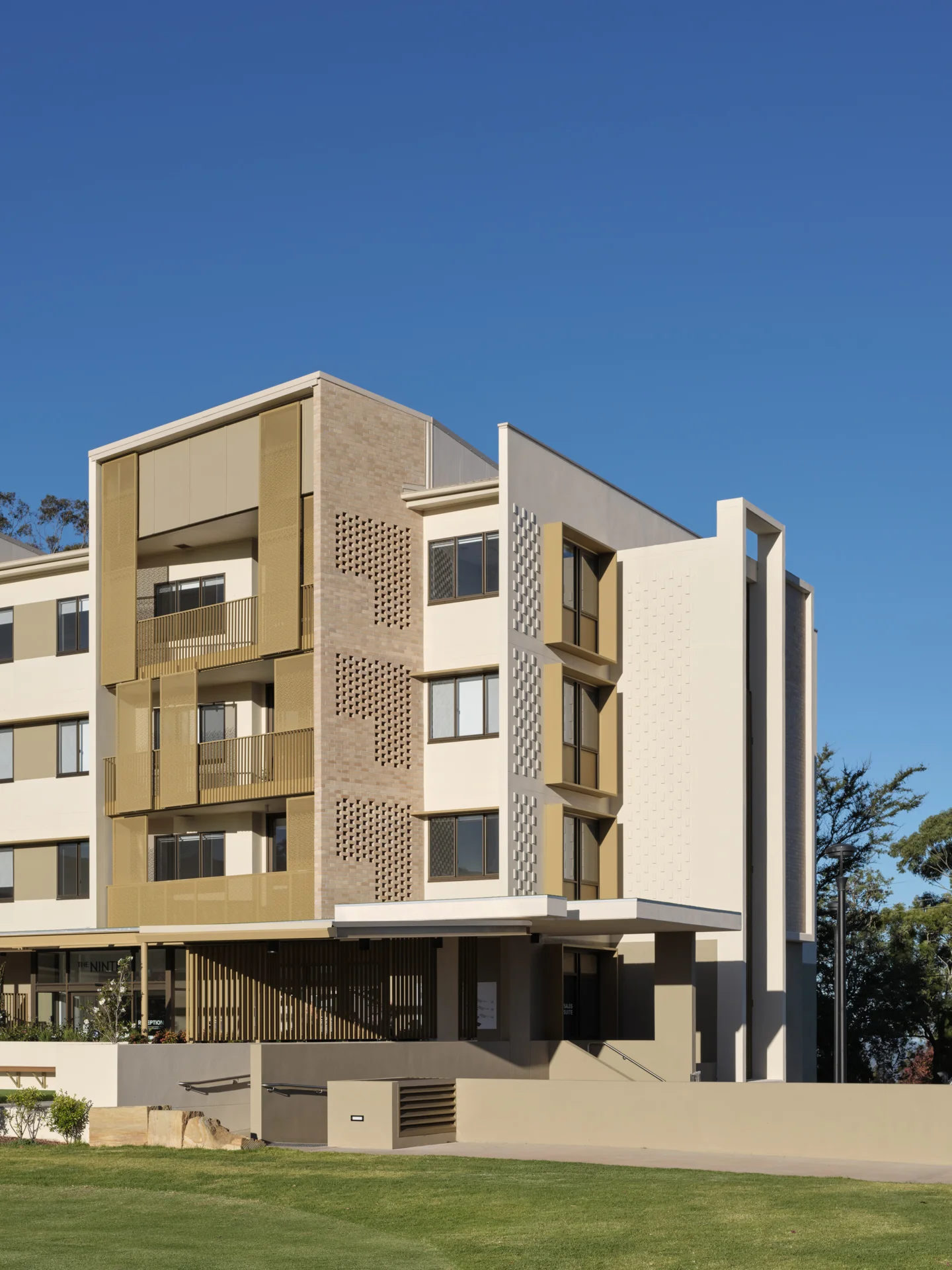 A modern, multi-story apartment building with beige and white exterior panels, geometric architectural details, and balconies, set against a clear blue sky and surrounded by a grassy lawn.