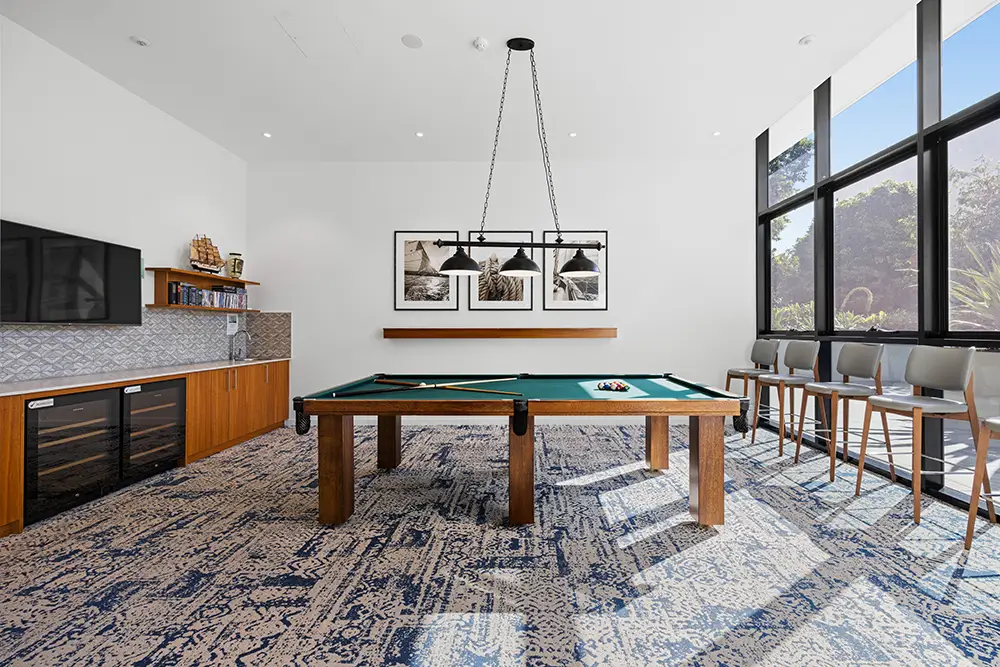 Modern game room with a pool table at center, cue rack and balls on the table, bar stools by large floor-to-ceiling windows, wall art, built-in cabinets with a TV, shelves, and wine coolers on the left.