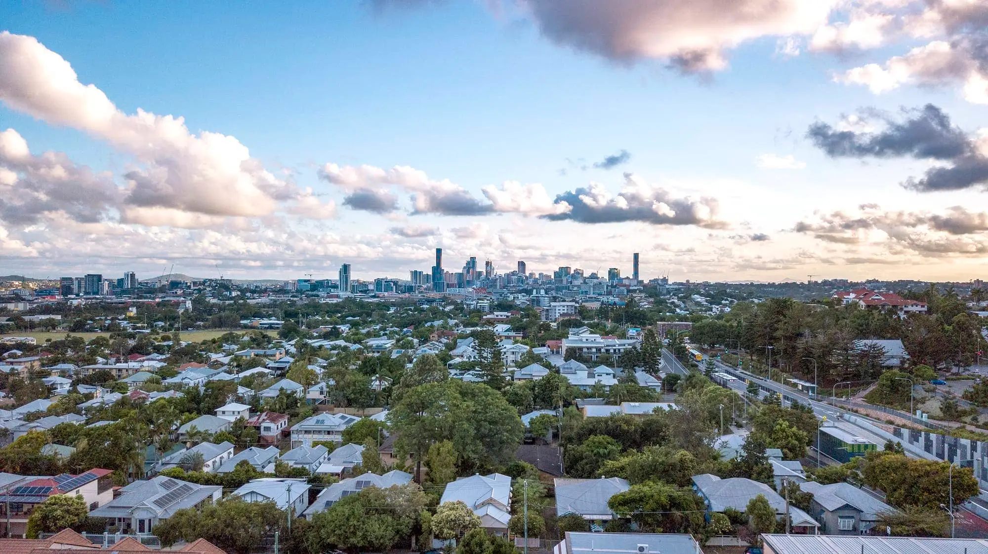 Aerial view of a suburban neighborhood with many houses and trees in the foreground, and a city skyline with tall buildings in the background under a partly cloudy sky.