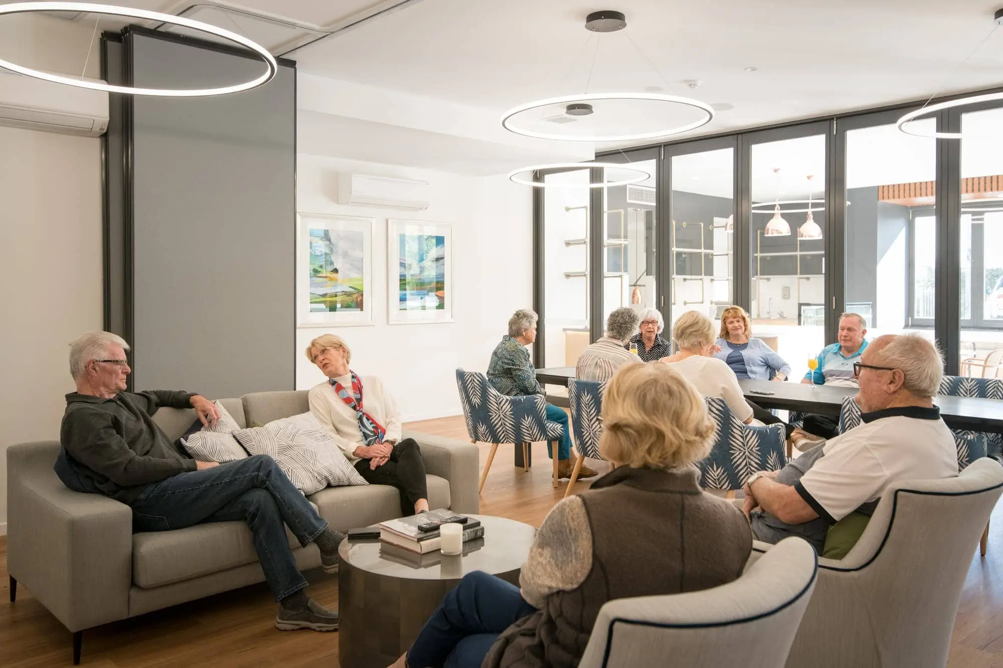 A group of seniors sit and chat in a bright, modern common room with large windows, contemporary furniture, and artwork on the walls. Some sit on sofas, others at a dining table, all engaged in conversation.
