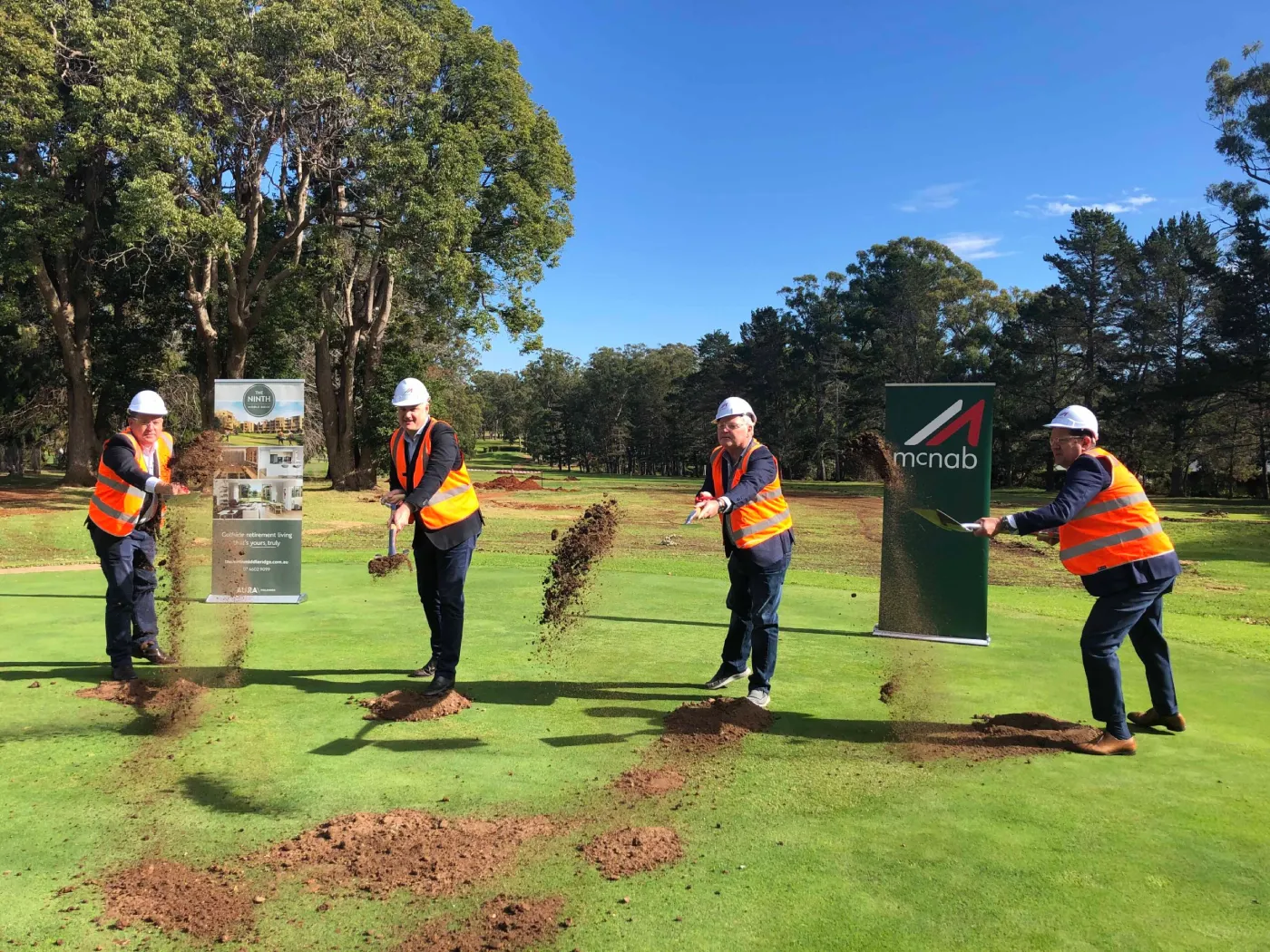 Four people in hard hats and orange safety vests toss dirt with shovels at a groundbreaking ceremony on a grassy area, with trees and two company banners in the background.
