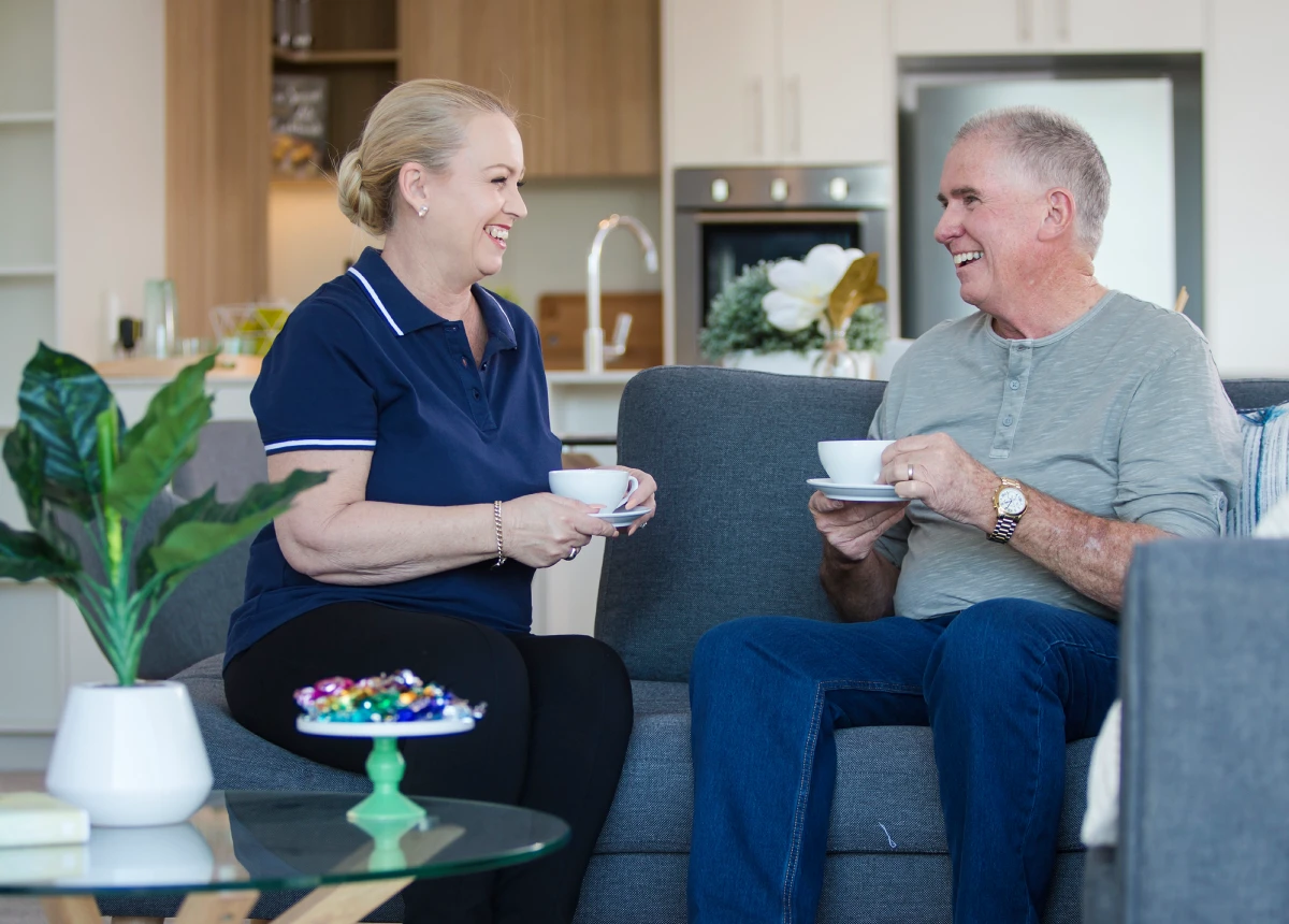 Two older adults sit on a couch in a modern living room, smiling and holding teacups. A dish of wrapped candies and a potted plant are on the coffee table in front of them. They appear to be enjoying a pleasant conversation.