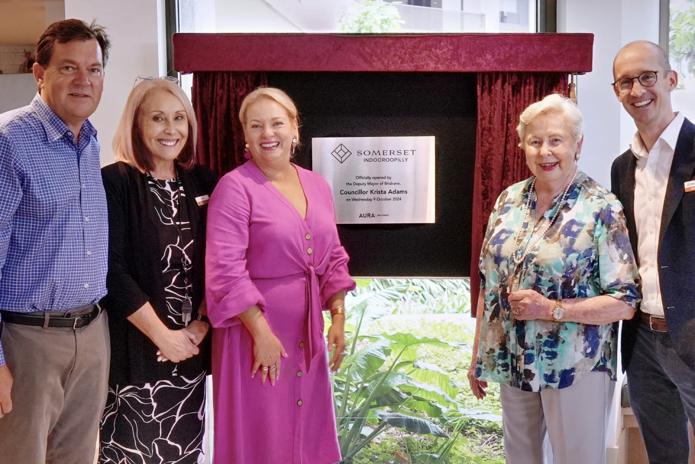 Five adults stand smiling next to a plaque for Somerset Indooroopilly’s official opening, partially covered by burgundy curtains. Large windows behind them show greenery outside.
