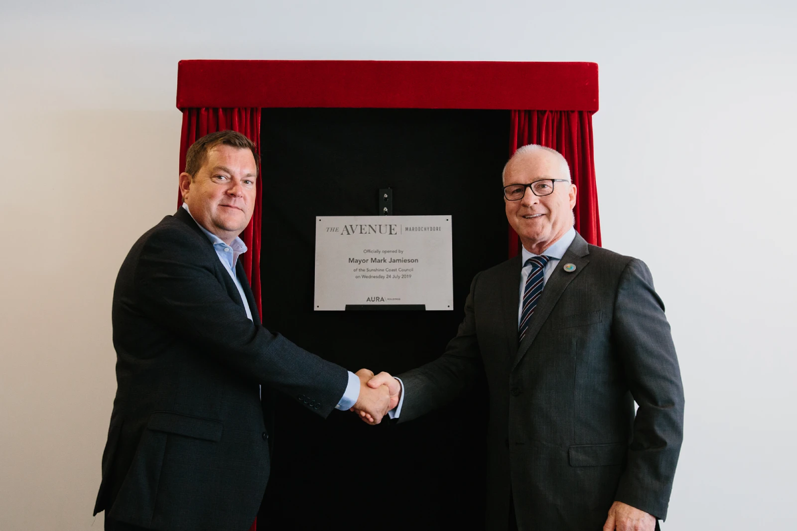 Two men in suits shake hands in front of a plaque mounted on a black stand with red curtains, suggesting the unveiling of a commemorative sign at an official event.