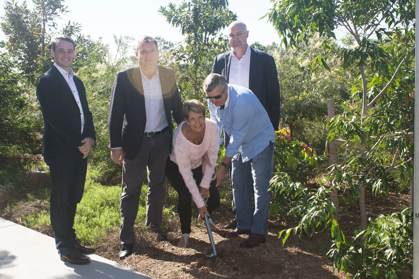 Five people, four men in suits and one woman in a pink sweater, participate in a groundbreaking ceremony outdoors in a garden area; two people hold a shovel together, surrounded by greenery and trees.
