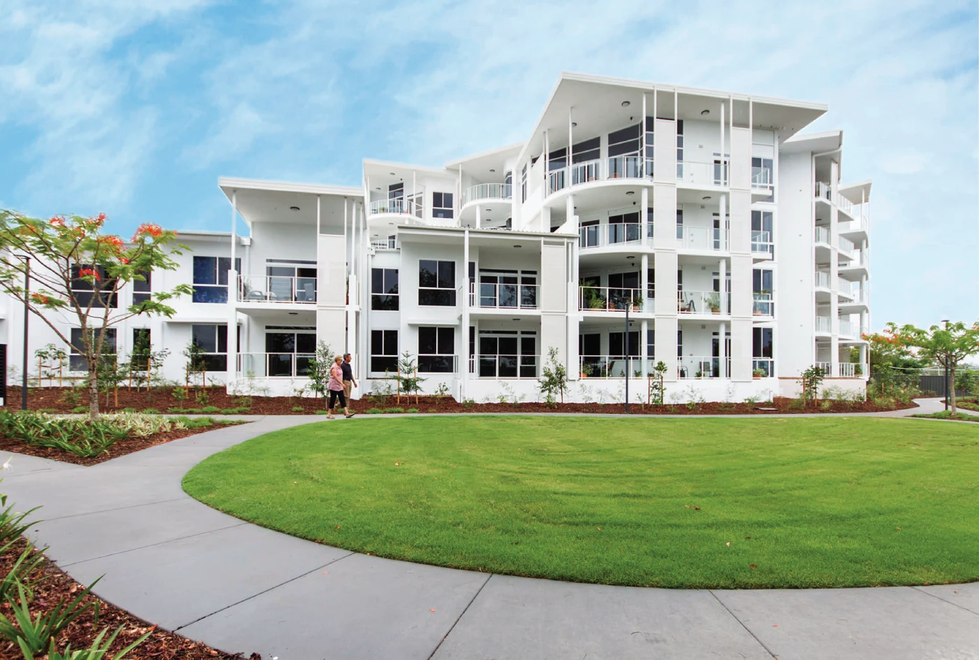 A modern white apartment building with multiple balconies stands behind a well-maintained green lawn and a curved paved walkway, with two people walking near the entrance under a blue sky.