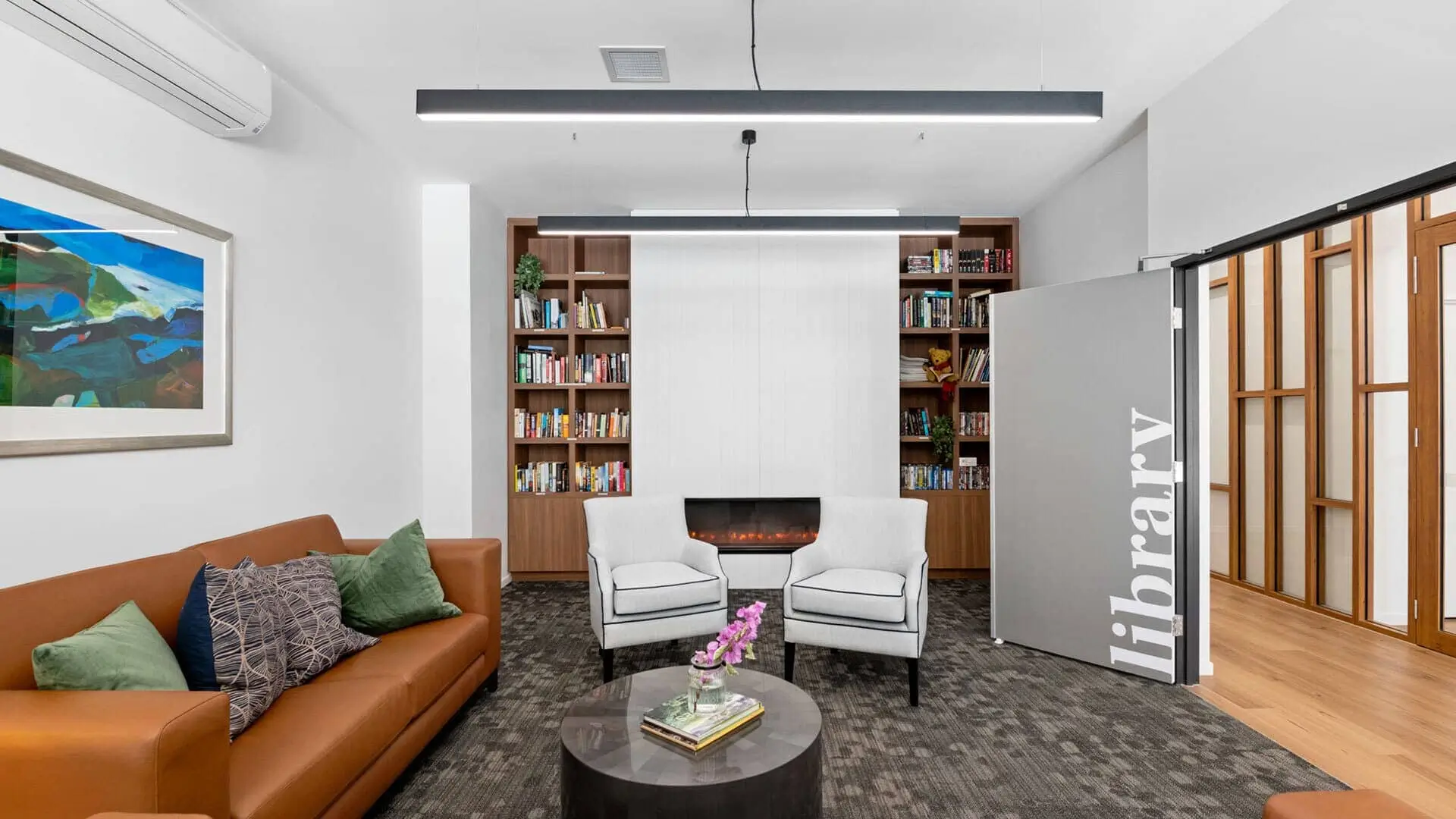 Modern library lounge with a brown sofa, two white armchairs, a round coffee table with flowers, bookshelves, and a partially open door labeled "library." Artwork and green pillows add color to the cozy, well-lit space.