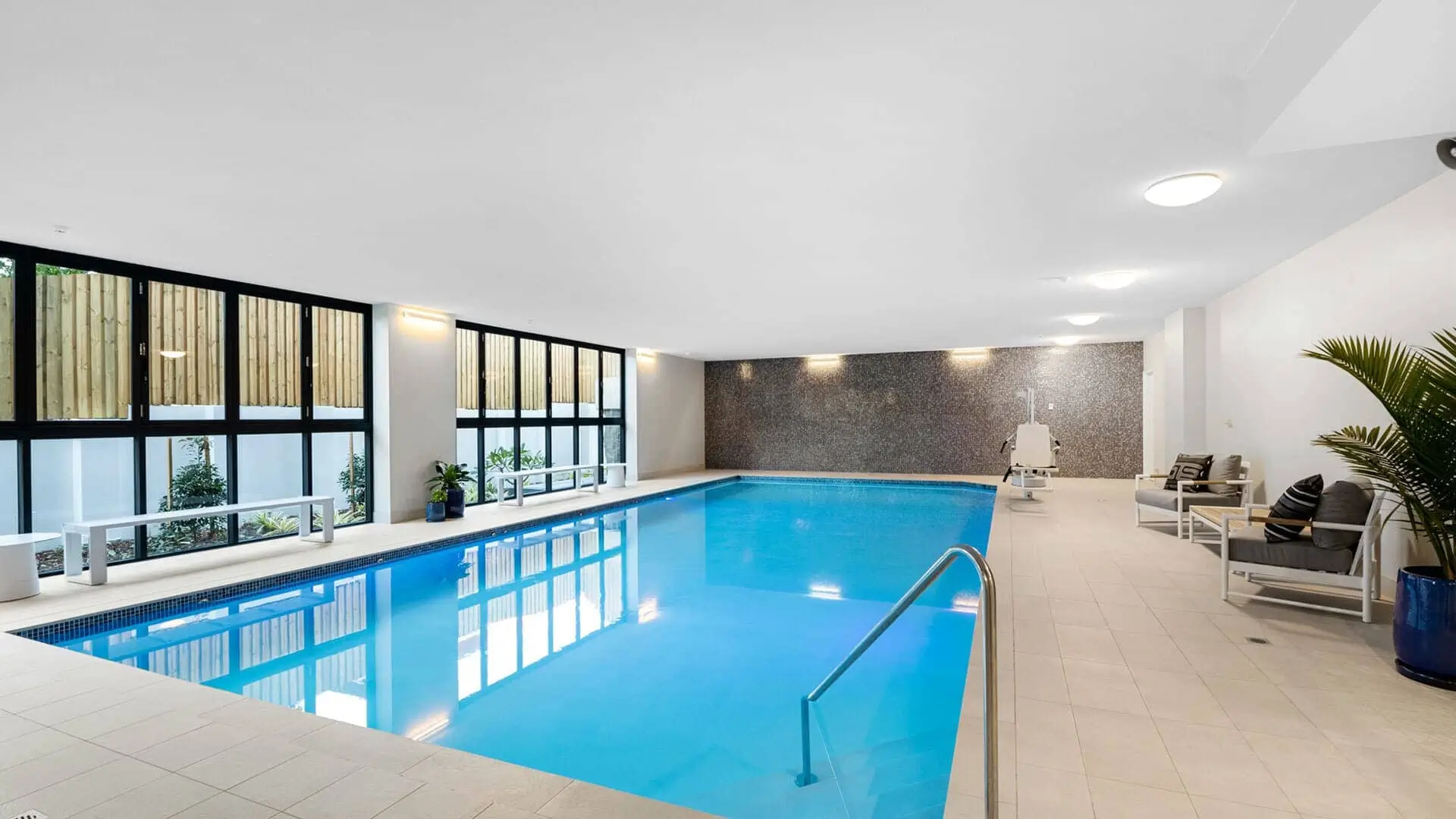 An indoor swimming pool with clear blue water, surrounded by beige tiles. Large windows line one wall, letting in natural light. There are lounge chairs, potted plants, and modern lighting around the pool area.