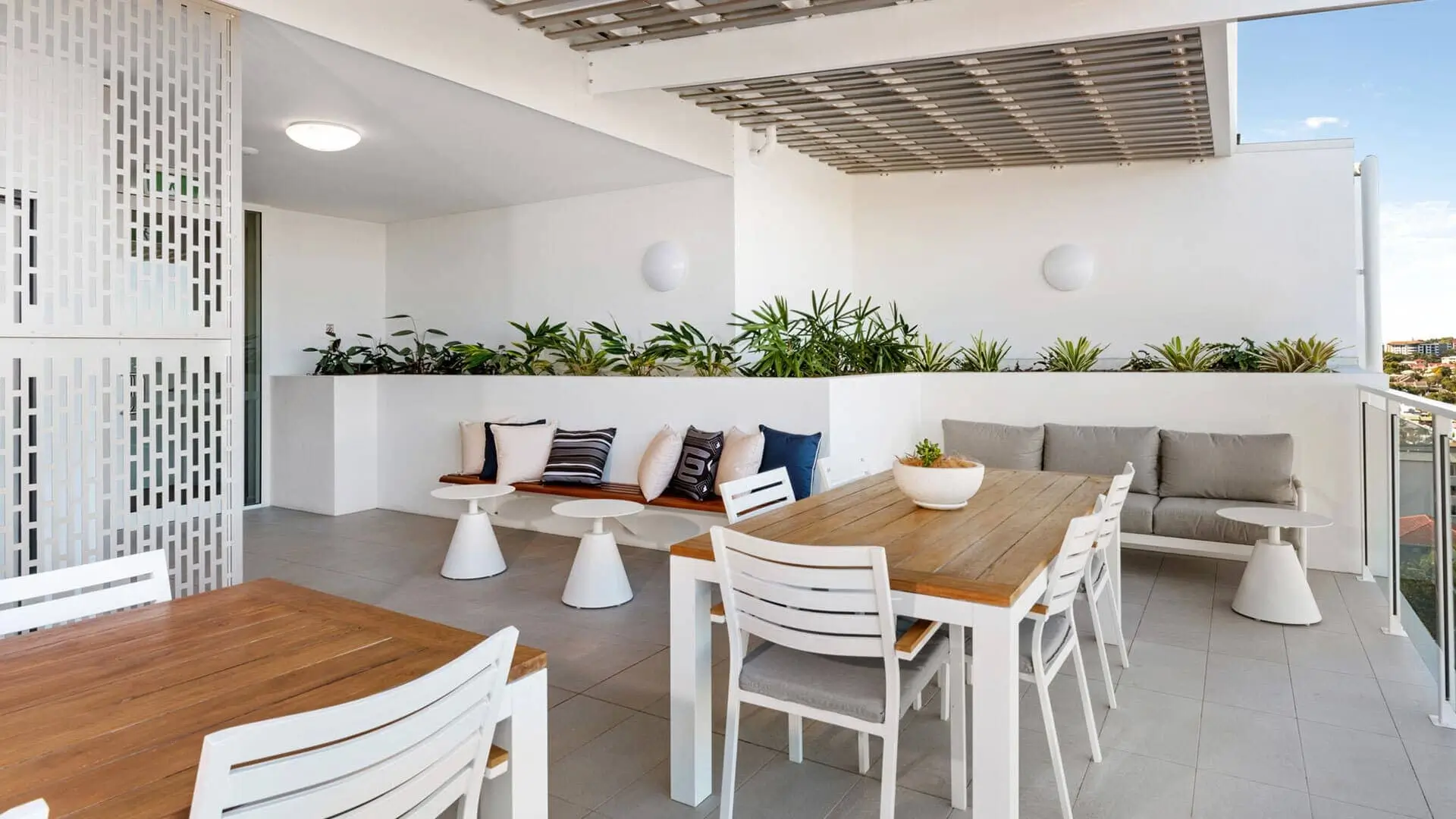 Modern outdoor patio with white walls and tiled floor, featuring wooden dining tables, white chairs, a bench with colorful cushions, small tables, and a row of green plants along a planter. Bright and spacious setting.