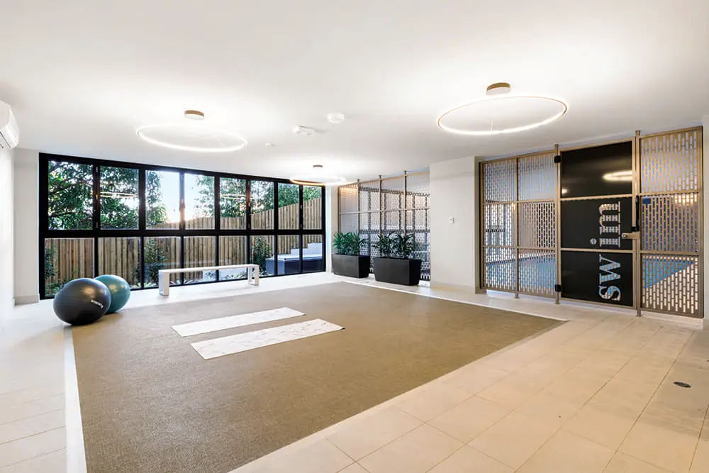 A bright, spacious exercise room with large windows, two yoga mats, two exercise balls, potted plants, and a bench. The room features modern lighting and a partition with the word "SWIM" partially visible.