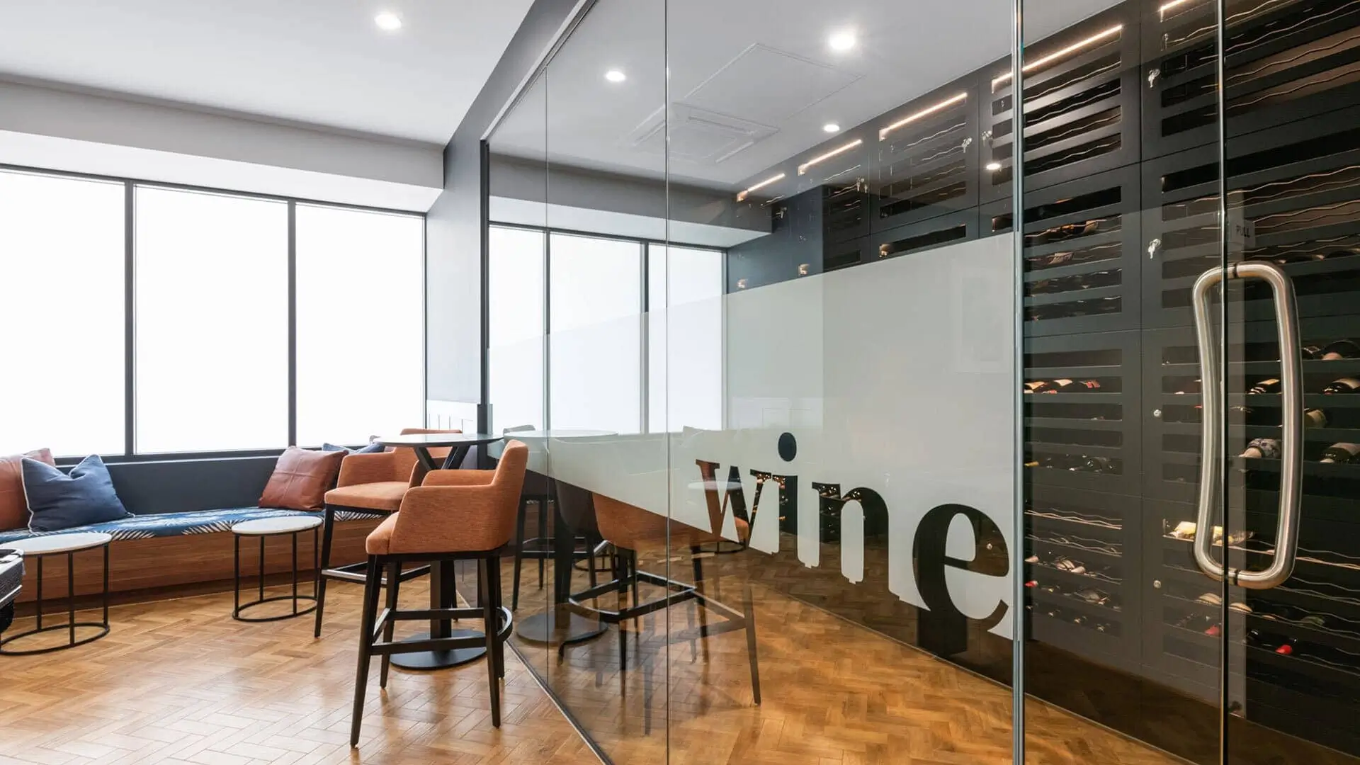 A modern office lounge with orange chairs, a sofa, and a glass-walled wine room labeled "wine." The space has large windows, wood flooring, and wine racks visible through the glass wall.