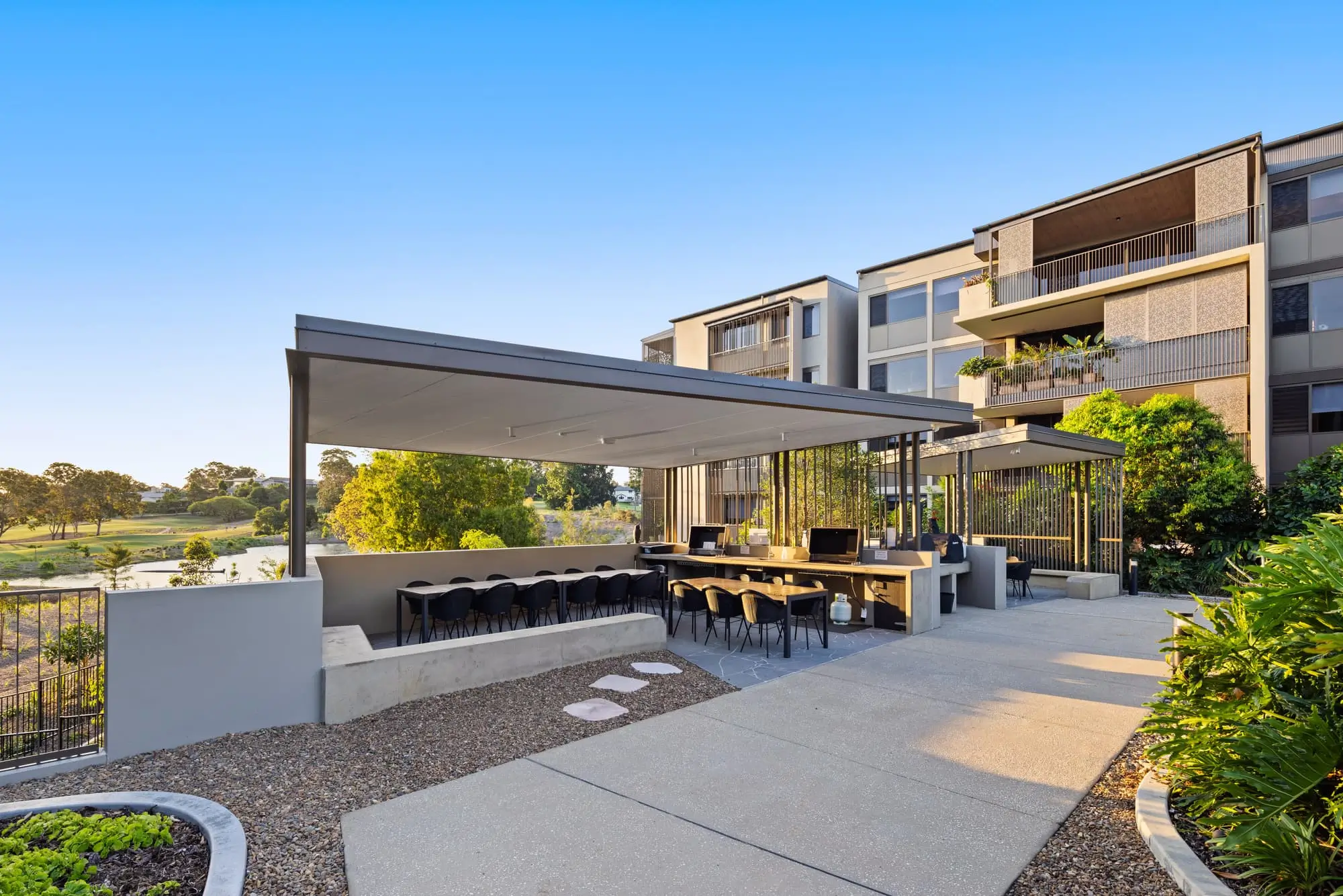A modern outdoor covered seating area with tables and chairs is set beside a multi-story apartment building, surrounded by greenery and landscaping under a clear blue sky.