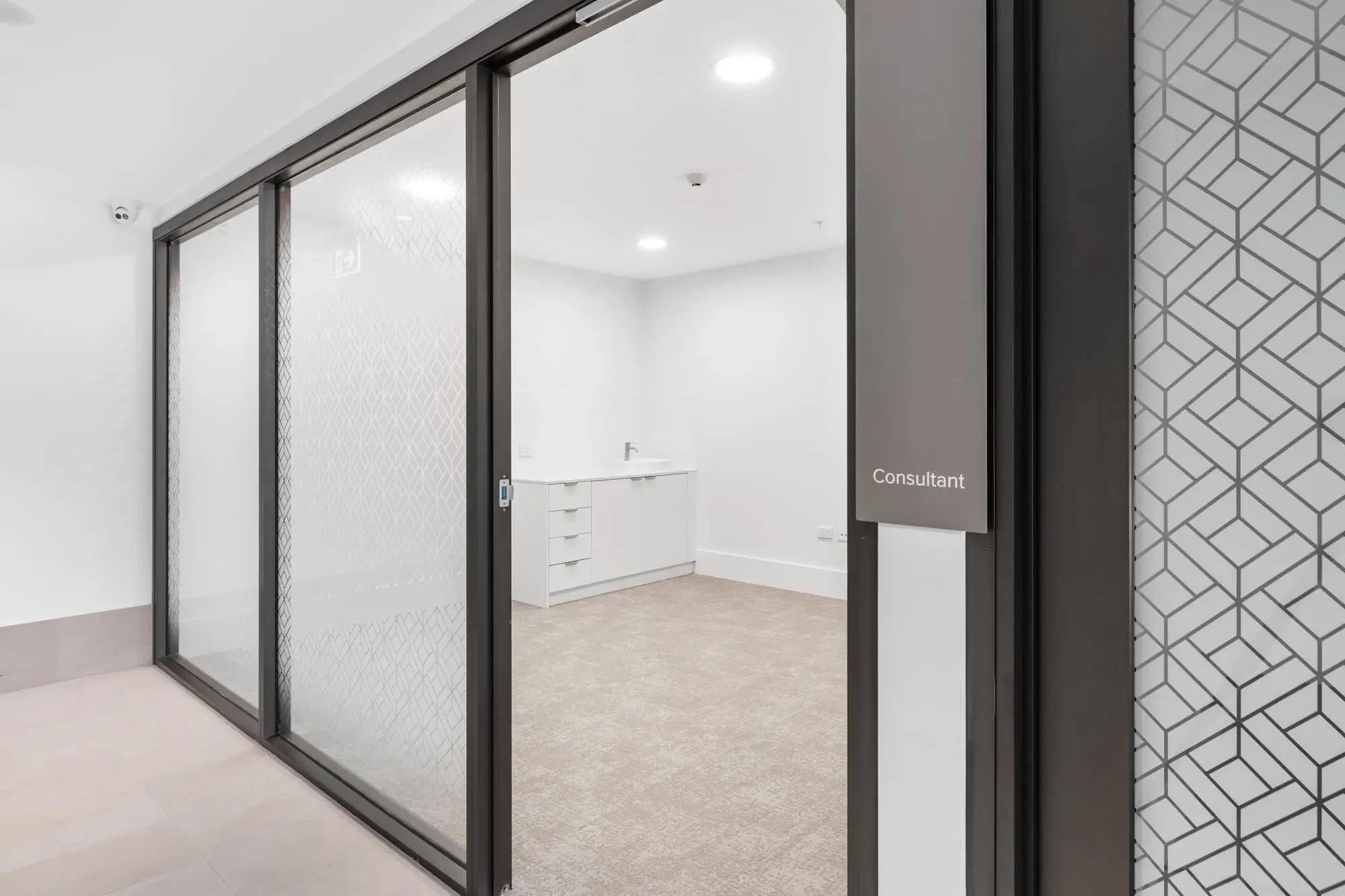 A modern, empty consultation room with frosted glass sliding doors, beige carpet, and white walls. A small sink with cabinets is visible, and a sign on the wall reads "Consultant.