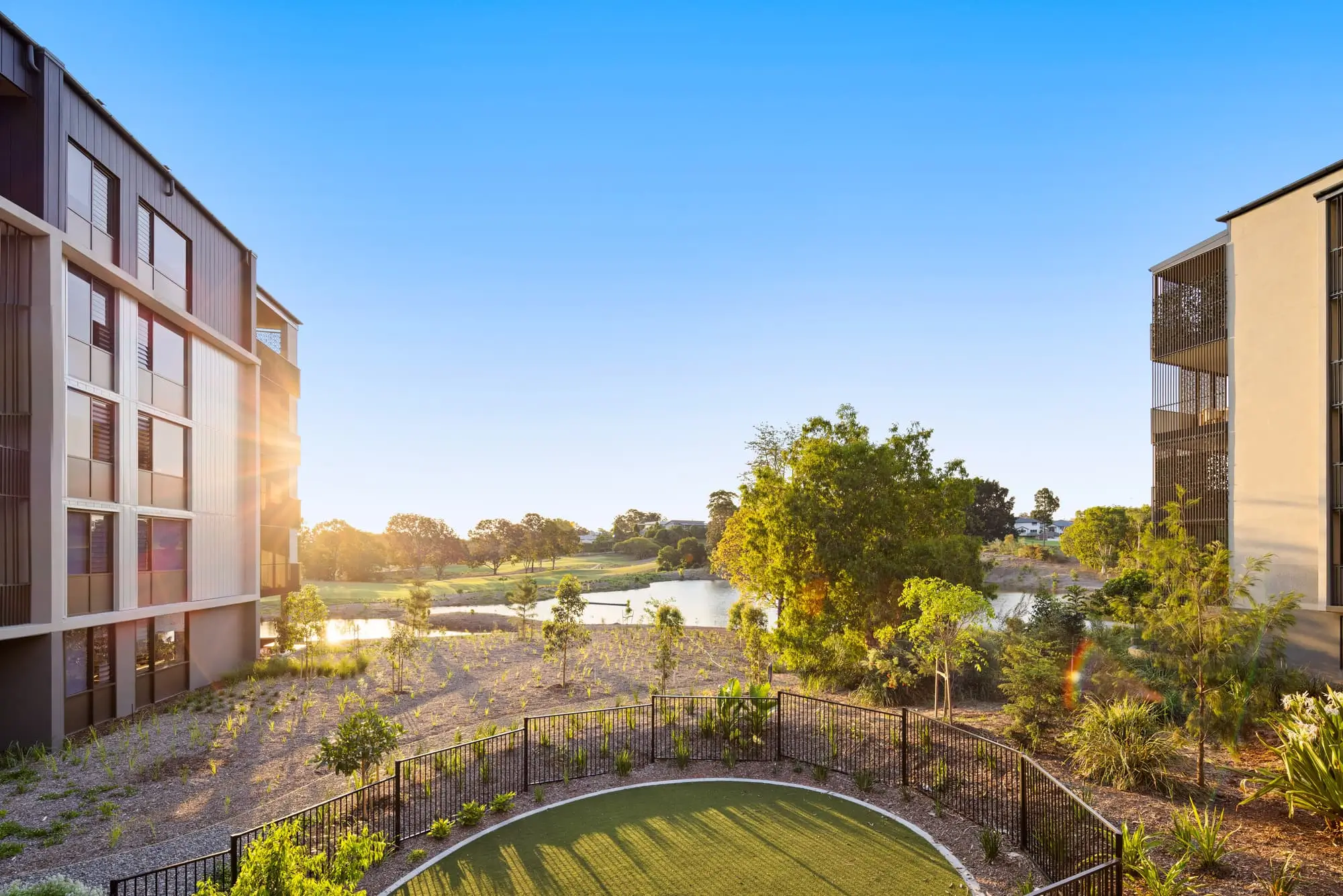 View of landscaped gardens and a small green lawn area with a black fence between two modern apartment buildings, overlooking a pond and trees under a clear blue sky at sunset.