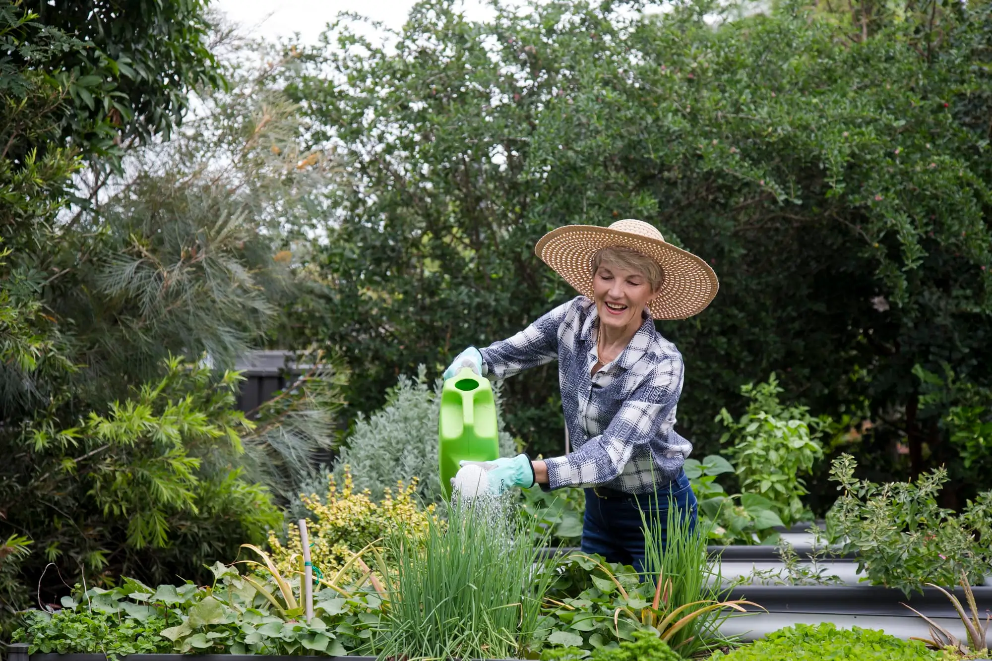 A woman wearing a straw hat, plaid shirt, and gloves smiles while watering plants with a green watering can in a lush garden. Various leafy plants and greenery surround her.