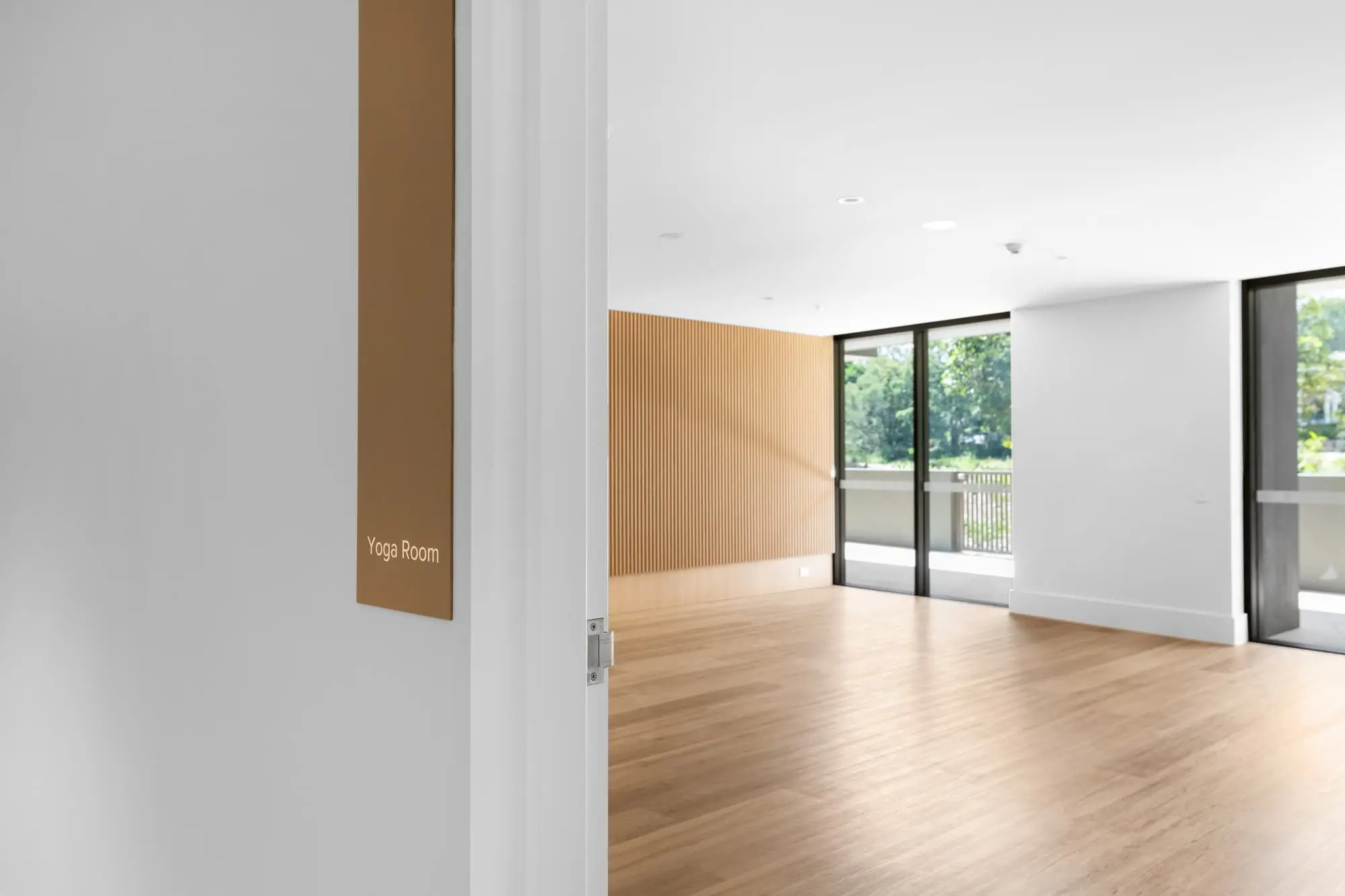 A bright, empty room with wooden floors and large windows is labeled "Yoga Room" on a sign next to the door. Sunlight streams in, and greenery is visible outside through the windows.