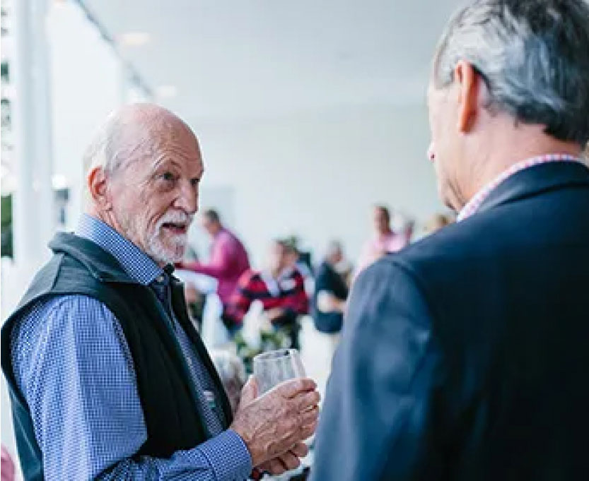 An older man holding a glass talks to another man in a suit at an outdoor gathering, with blurred people in the background.