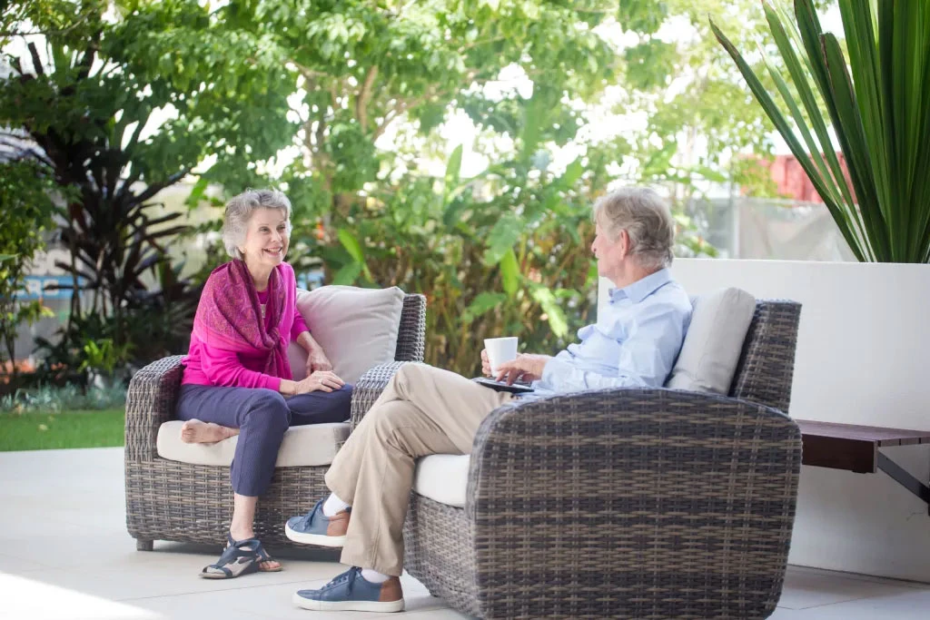 Two older adults sit on wicker patio furniture, smiling and talking together outdoors. Green plants and trees surround the bright, peaceful setting. One person holds a cup, and both appear relaxed and engaged in conversation.