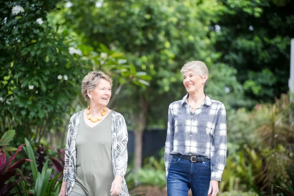 Two older women walk together outdoors in a lush garden, smiling and talking. One wears a patterned jacket over a light green outfit with a yellow necklace, the other wears a plaid shirt and jeans. Green trees and plants surround them.