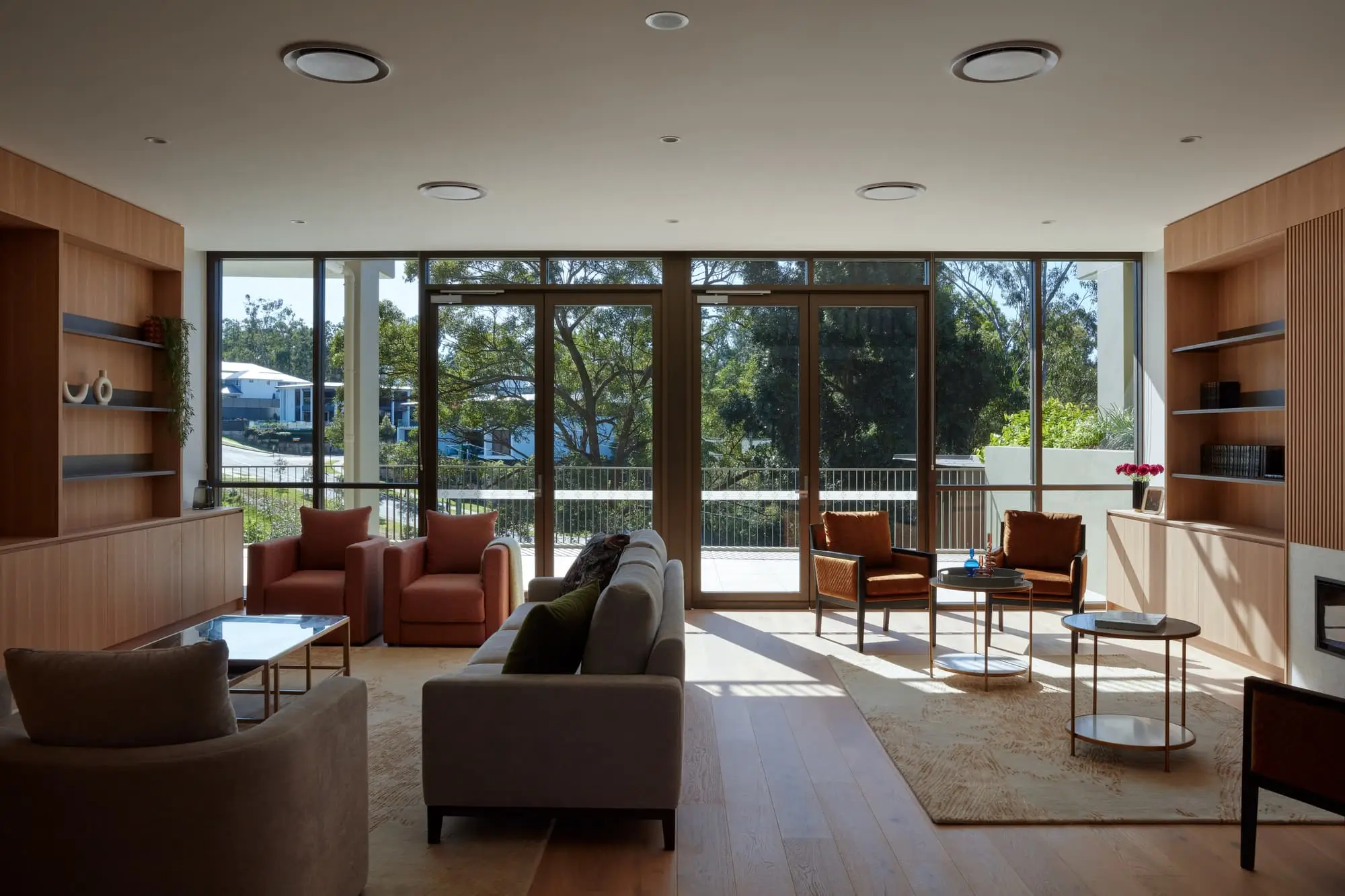 A modern living room with large floor-to-ceiling windows, neutral-toned sofas and armchairs, wooden floors, built-in shelving, and a view of trees and houses outside. Natural light fills the space.