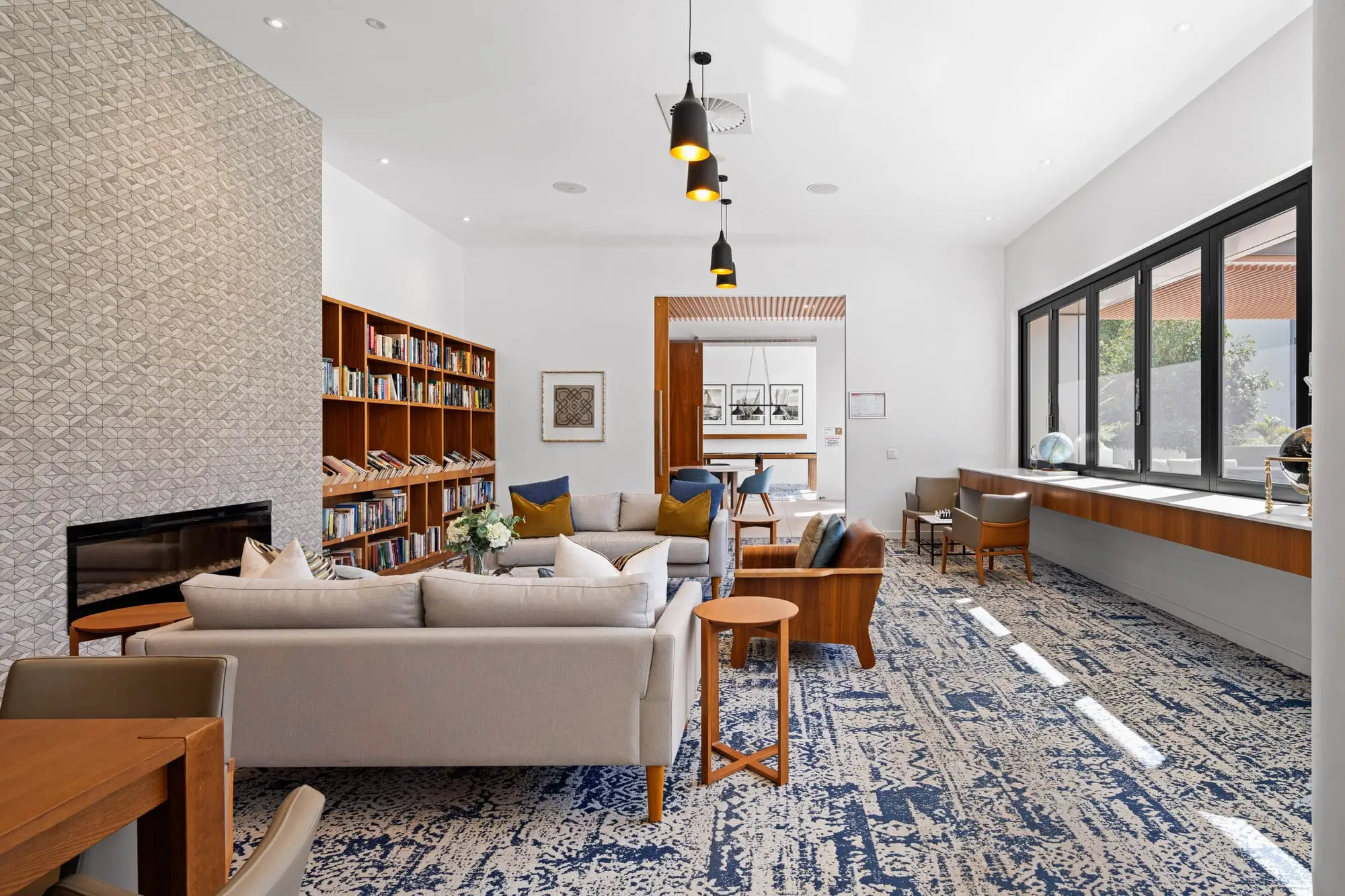 Modern, airy lounge with bookshelves, sofas, armchairs, and patterned blue carpet. Large windows let in natural light; a long desk runs below them. Contemporary light fixtures hang from the white ceiling.
