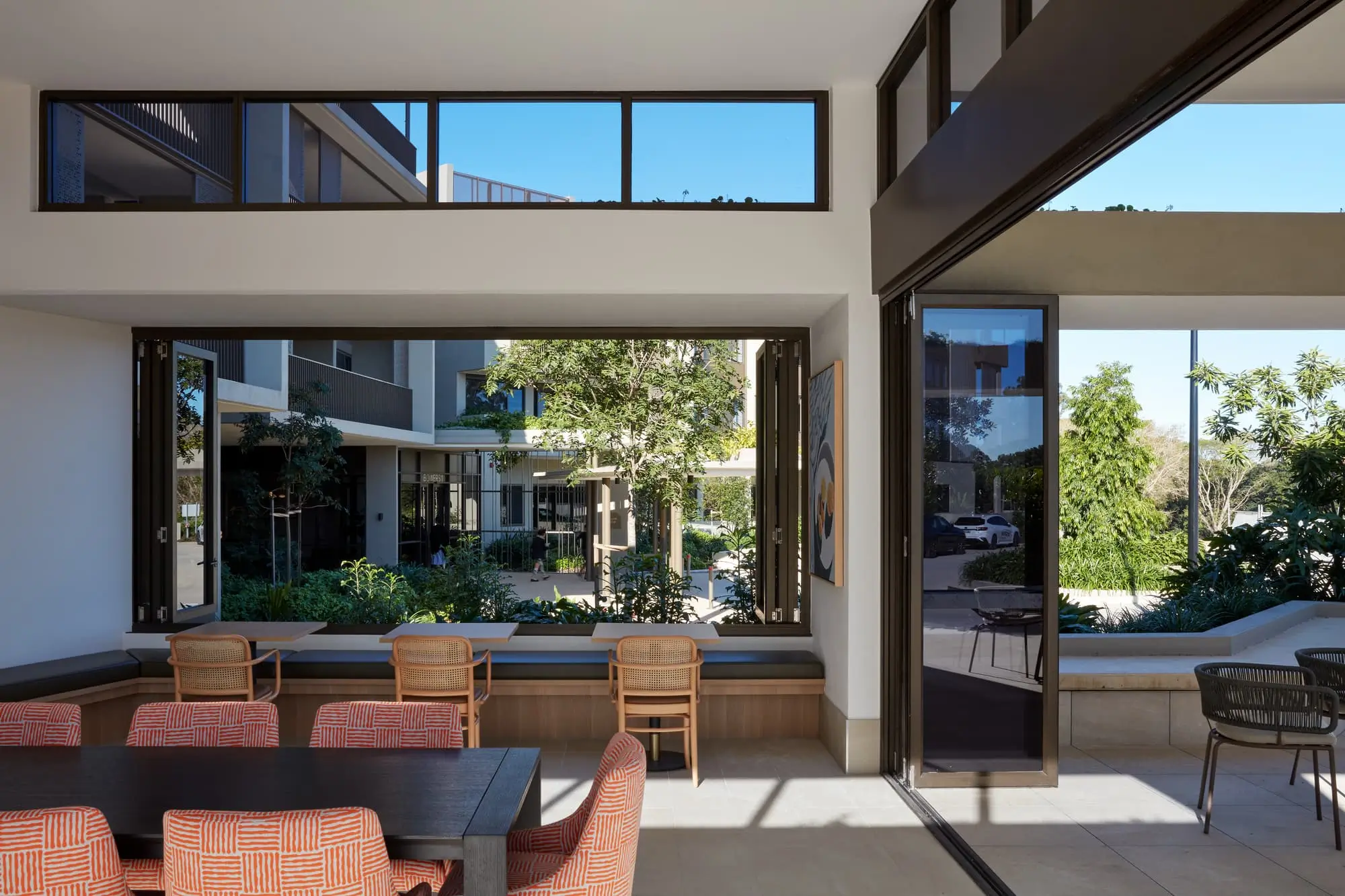 Modern indoor dining area with a black table, patterned chairs, and large open windows offering views of a lush, green courtyard and surrounding contemporary buildings. Bright natural light fills the space.