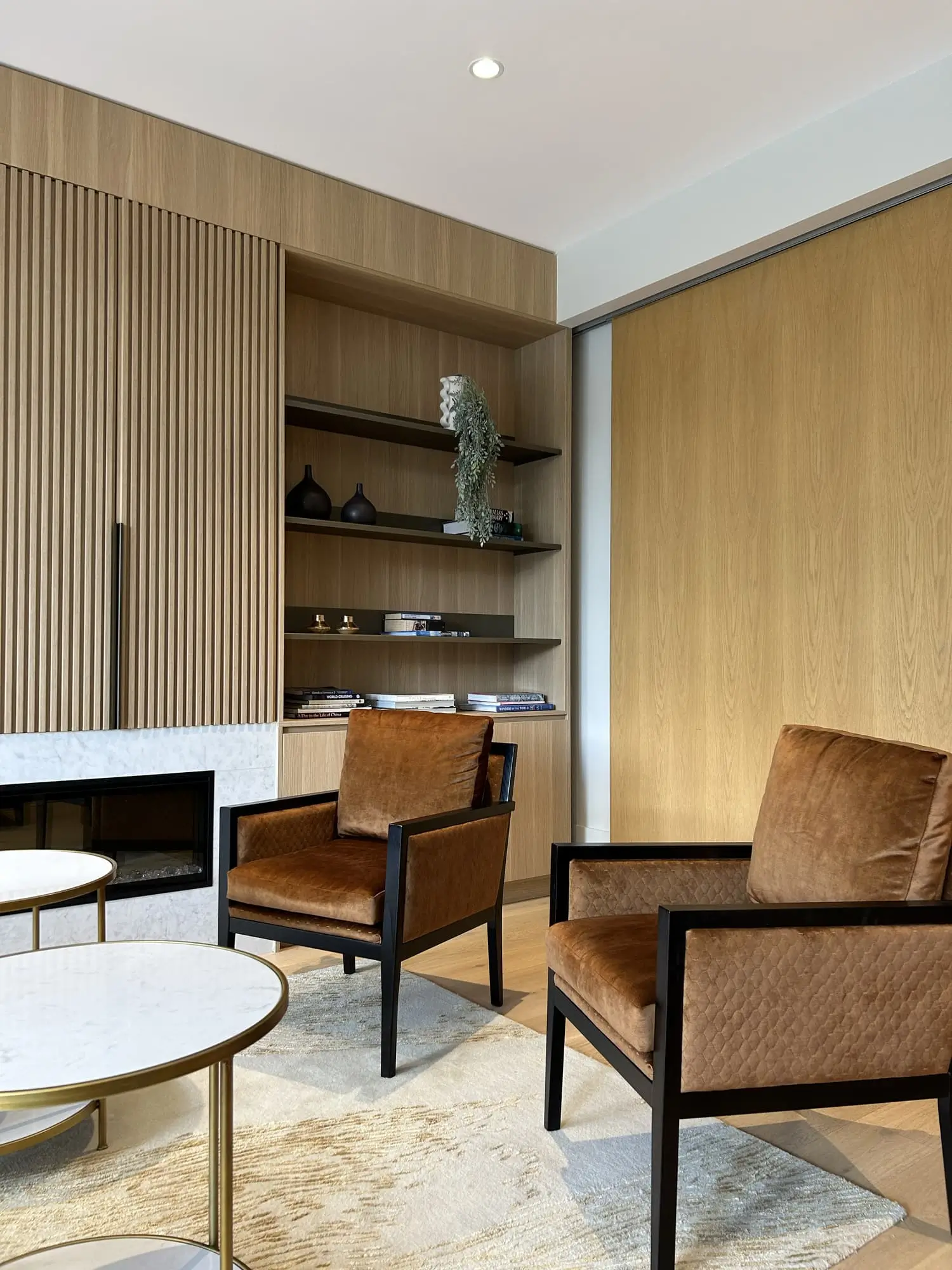 A modern living room with two brown leather armchairs, a light rug, marble-topped tables, and wood-paneled walls. Built-in shelves display books, vases, and a decorative sculpture.