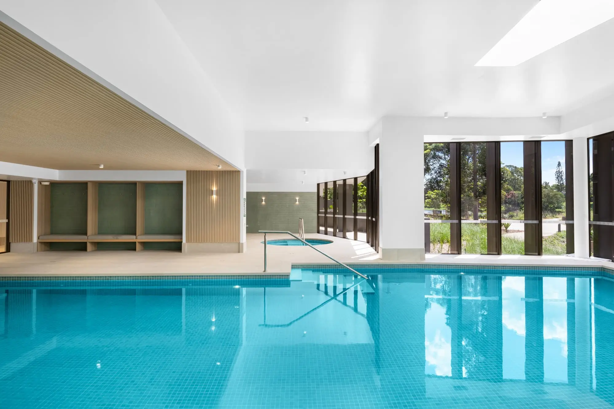 Indoor swimming pool with clear blue water, a handrail leading into the pool, wooden benches along one wall, and large floor-to-ceiling windows letting in natural light and showing greenery outside.