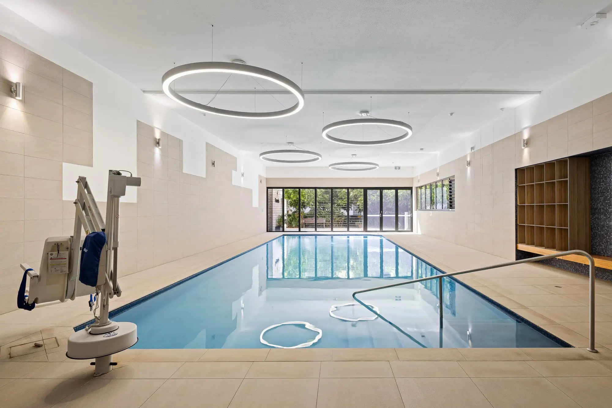 Modern indoor swimming pool with beige tiled walls and floor, circular overhead lights, a pool lift for accessibility, wooden cubby shelves, a bench, and large windows showing greenery outside.