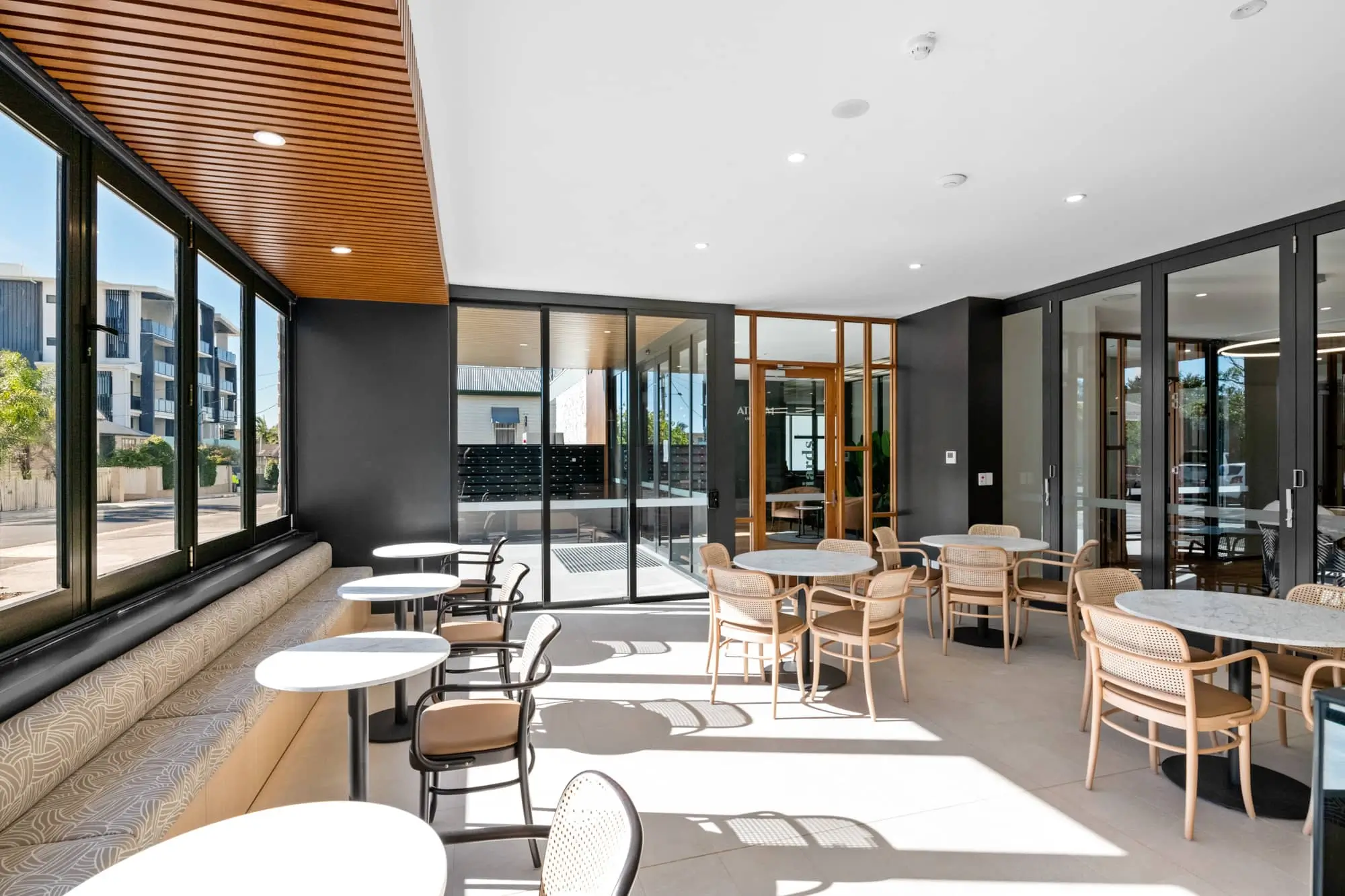 Modern cafe with round white tables and light wooden chairs next to large windows. Bright natural light fills the space, and glass doors lead to an outdoor patio. Neutral tones and clean lines create a welcoming atmosphere.