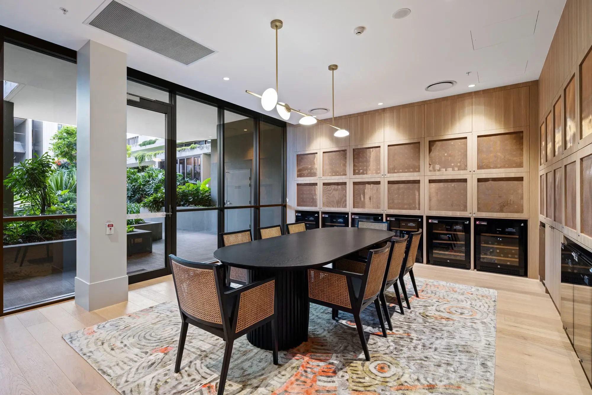 Modern meeting room with a black oval table, six woven chairs, built-in wooden wall shelves, a patterned rug, large glass doors, and greenery visible outside. Contemporary light fixture hangs above the table.