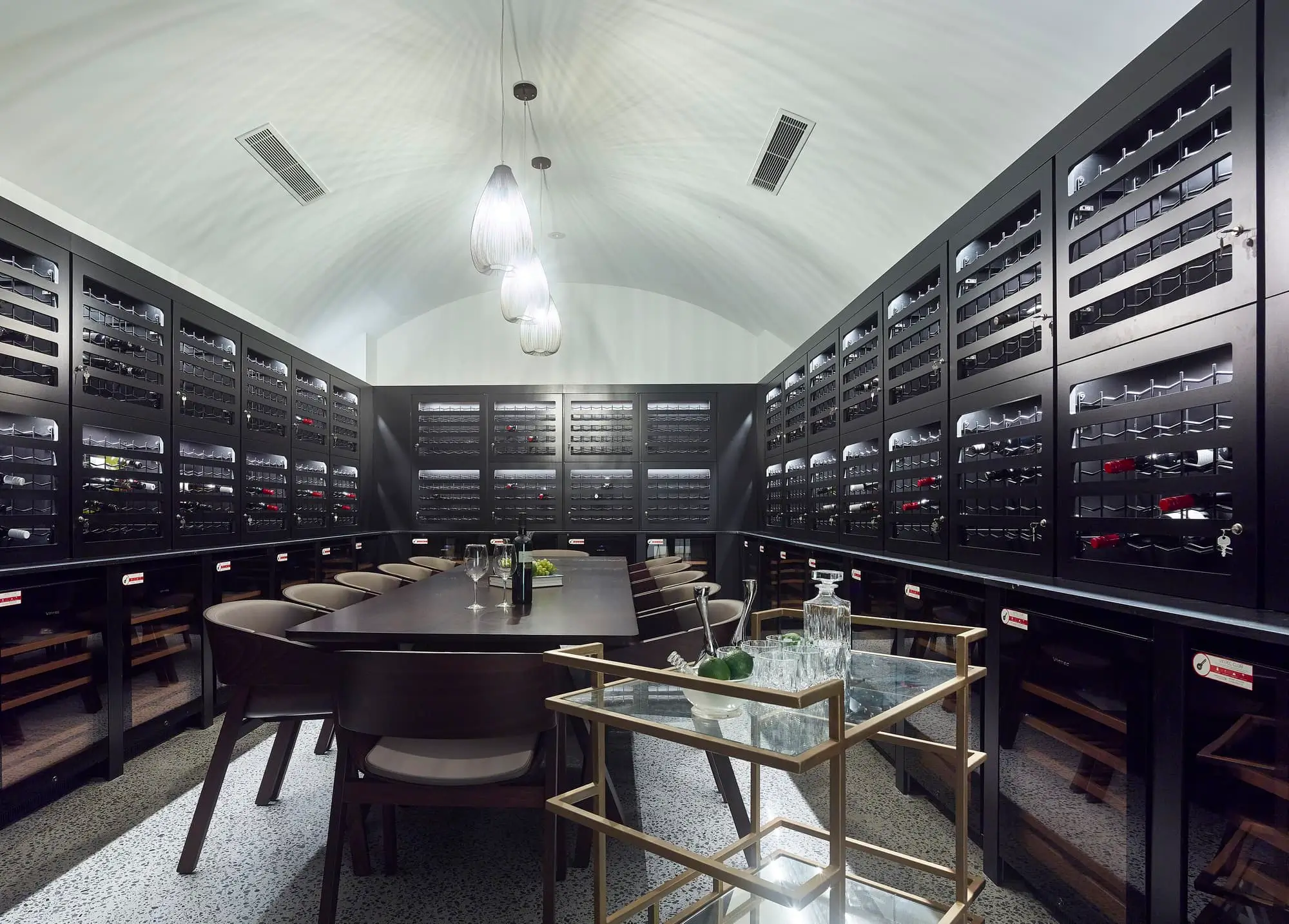 A modern wine cellar with black racks filled with wine bottles lines the walls. In the center is a long dining table with chairs, and a glass bar cart with bottles and glasses is positioned nearby under pendant lights.
