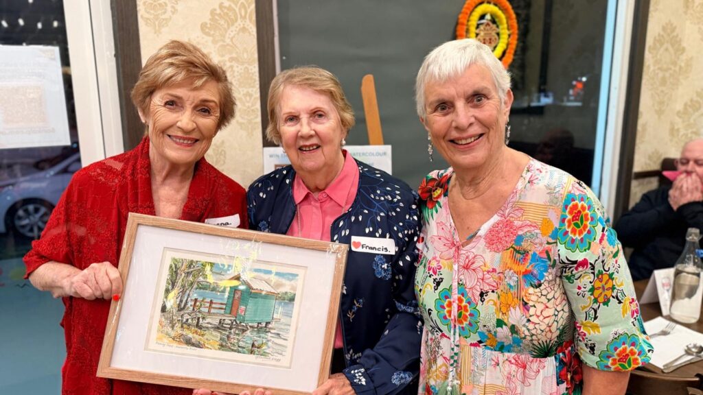 Three smiling older women stand together indoors; one holds a framed painting of a house by the water. Dressed in colorful clothing, they appear happy at this award-winning Somerset event, with other people and decorations visible in the background.
