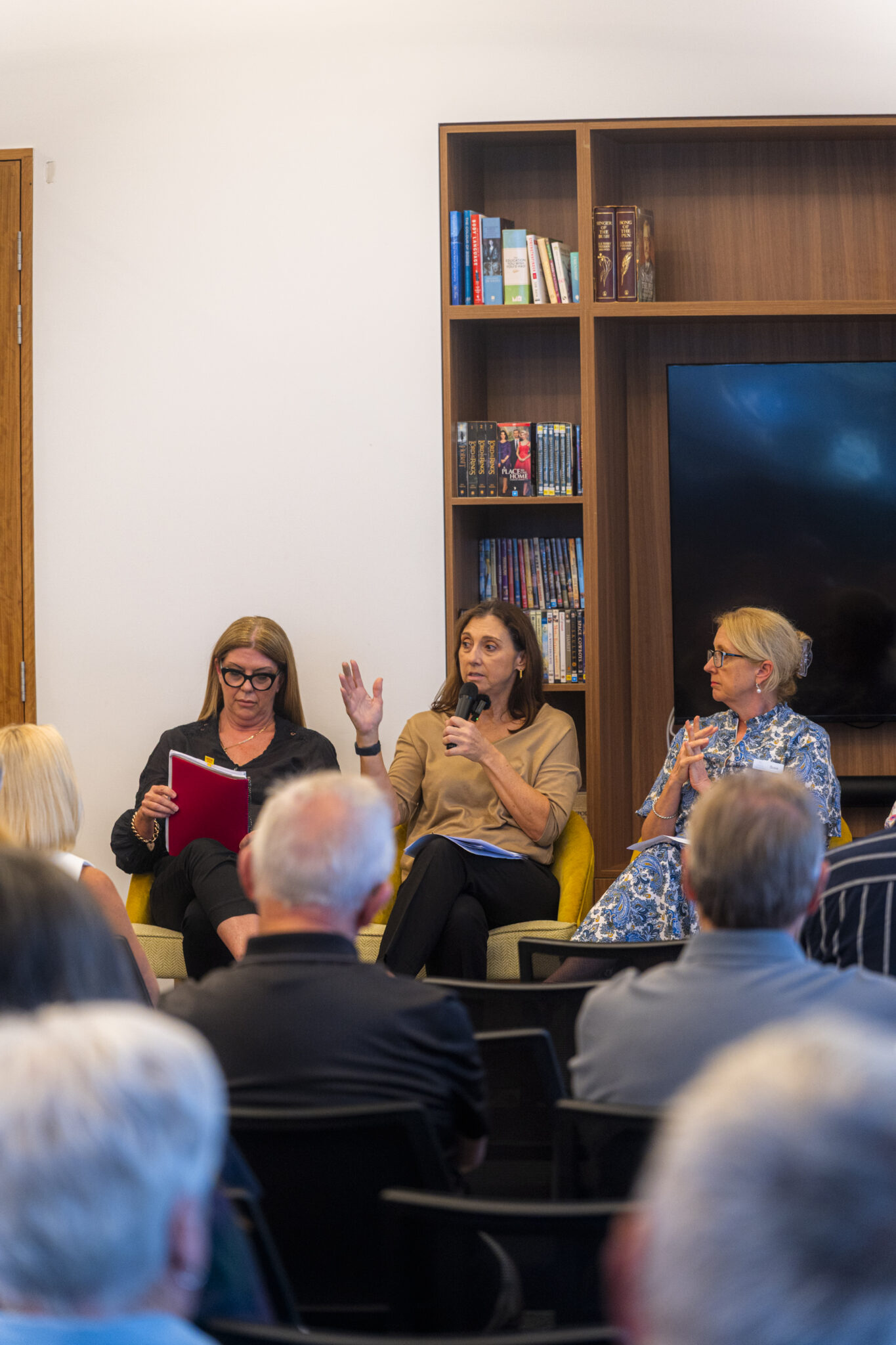 Three women sit on a panel in front of an audience; one holds a microphone and speaks, another holds notes and smiles, while the third woman listens. They are seated by shelves with books and DVDs.