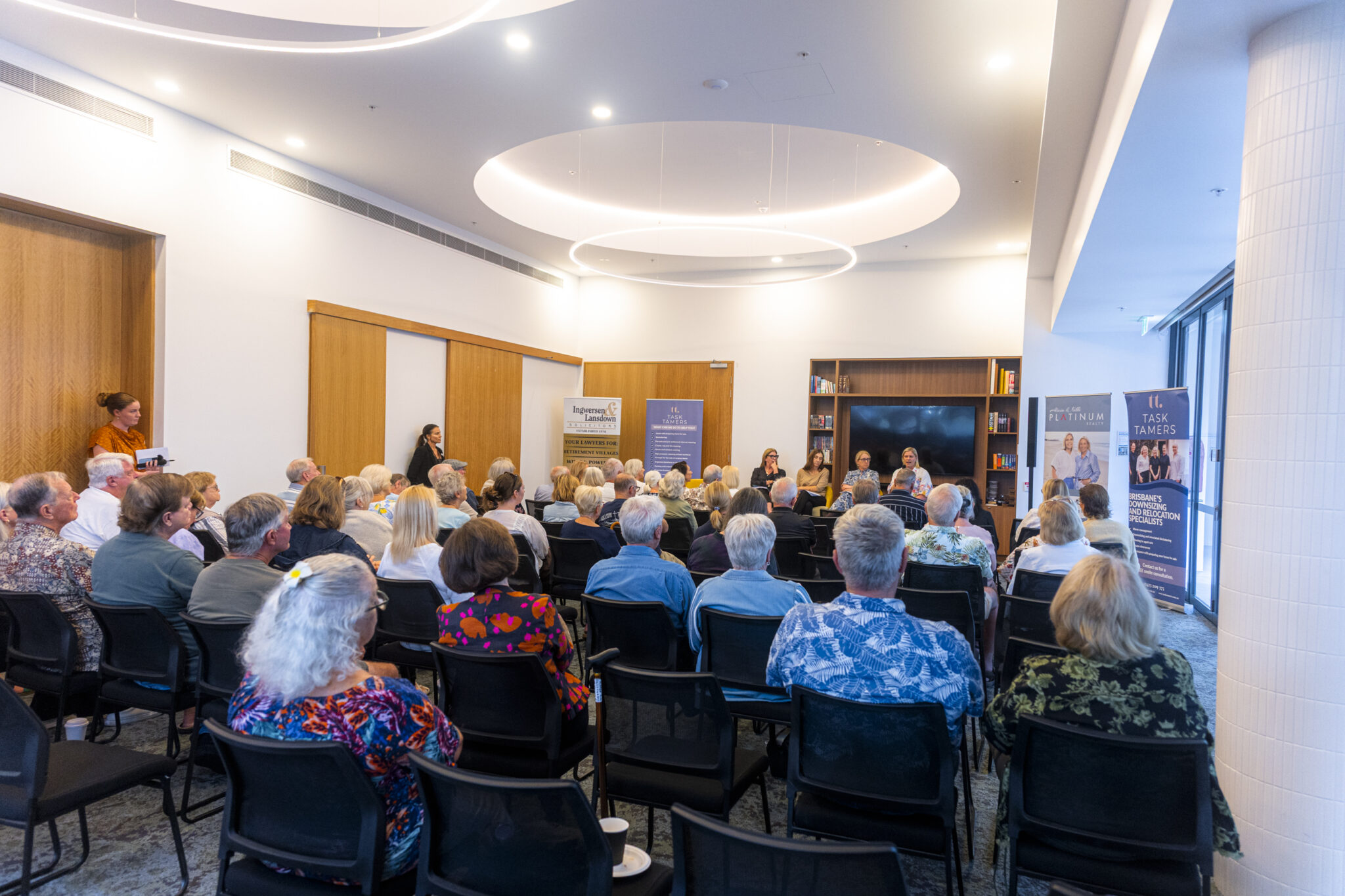 A group of people seated in a bright room listen to a panel discussion. Four speakers sit at the front with banners behind them. Most attendees have gray or white hair, and the atmosphere appears attentive and engaged.