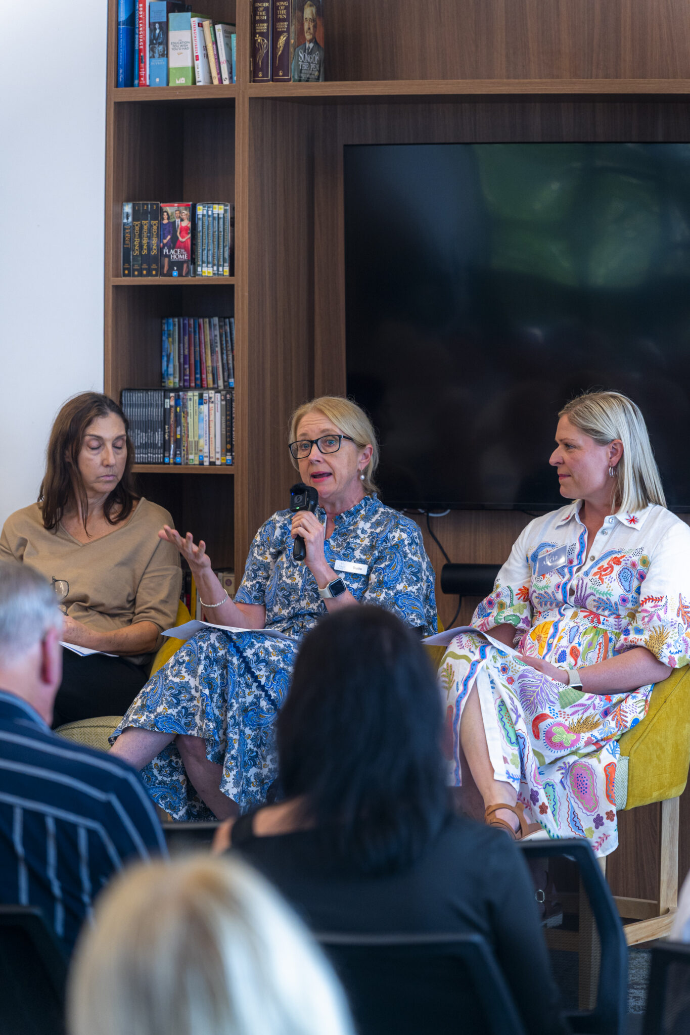 Three women sit in front of an audience, with one speaking into a microphone. They are seated by a bookshelf and a large TV screen, participating in what appears to be a panel discussion or event.