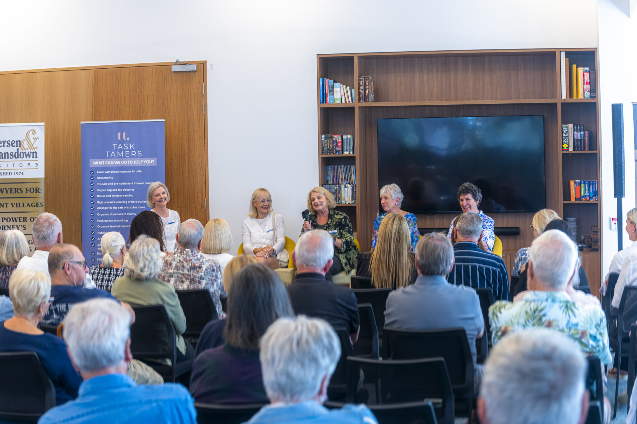 A panel of five women sit speaking in front of an audience in a library or meeting room, with bookshelves, a large TV, and banners in the background. The audience listens attentively, facing the speakers.