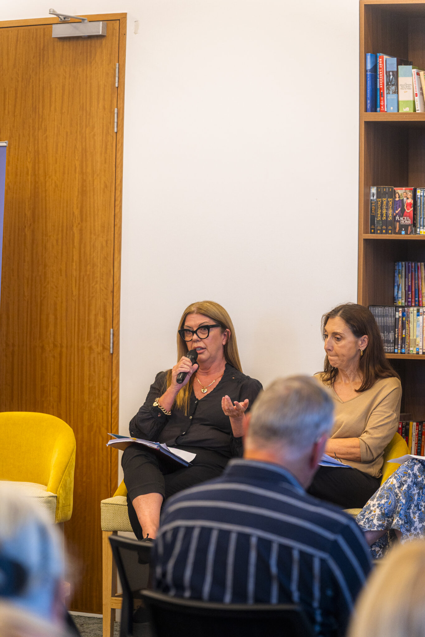 A woman holding a microphone speaks while seated next to another woman during an indoor event. People in the audience are visible in the foreground, and bookshelves are in the background.