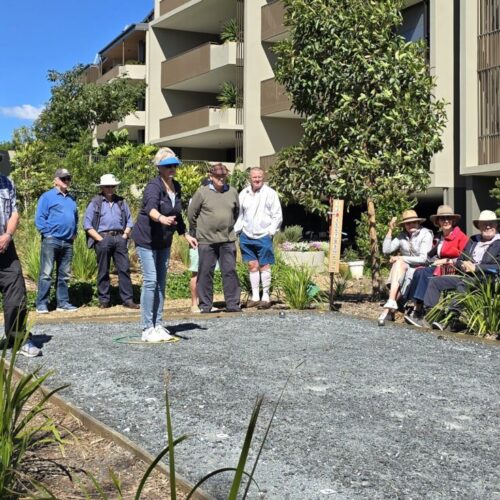 A group of people sit and stand outdoors on a sunny day, watching a man play pétanque on a gravel court, with modern apartment buildings and greenery in the background.
