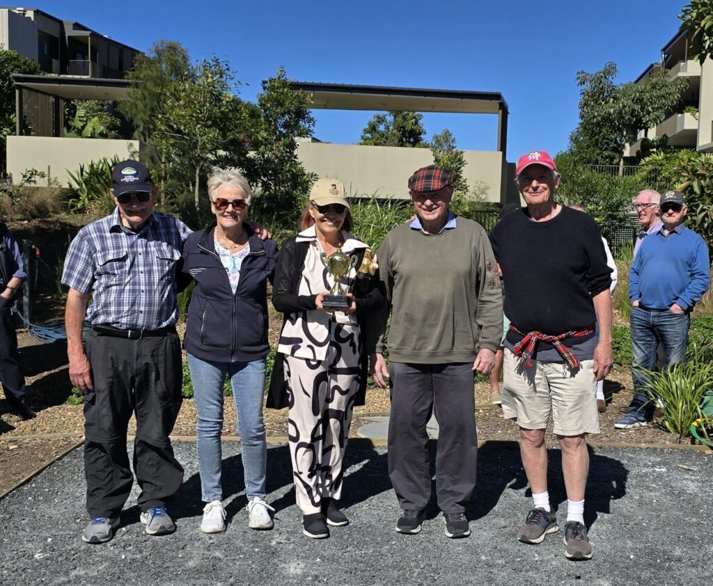 Five older adults stand outdoors in a row, smiling at the camera. The woman in the center holds a trophy. The group is casually dressed, and greenery and buildings are visible in the background under a clear sky.