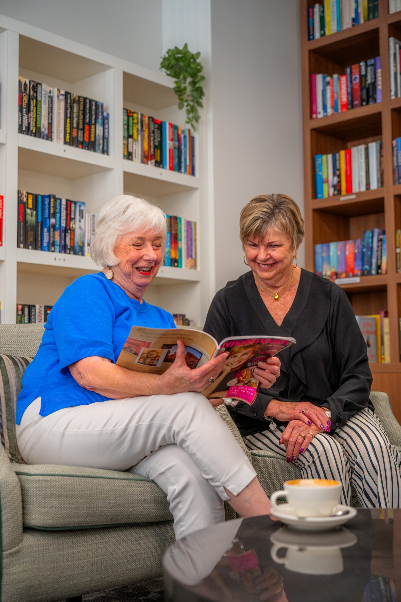 Two older women sit on a couch in a cozy library, smiling and looking at a magazine together. Bookshelves filled with colorful books are in the background, and a cup sits on the table in the foreground.