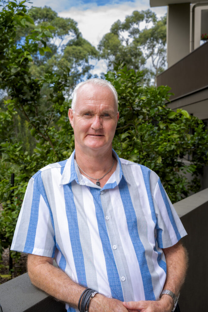 A middle-aged man with short gray hair wearing a blue and white striped shirt stands outside, leaning on a railing with trees and a building in the background.