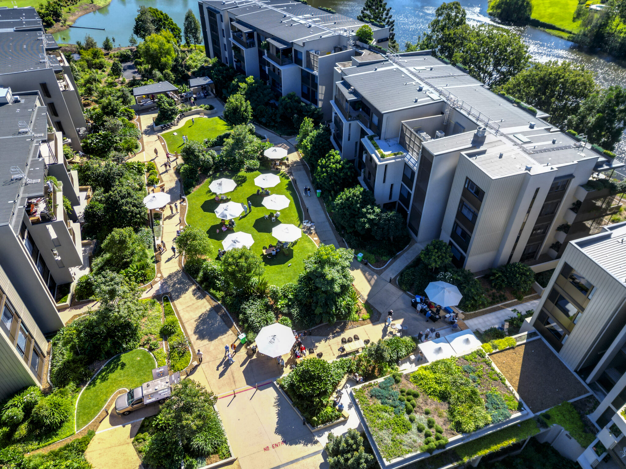 Aerial view of a modern apartment complex with green lawns, walking paths, trees, white patio umbrellas, and people socializing in the central courtyard; a pond and more greenery are visible in the background.